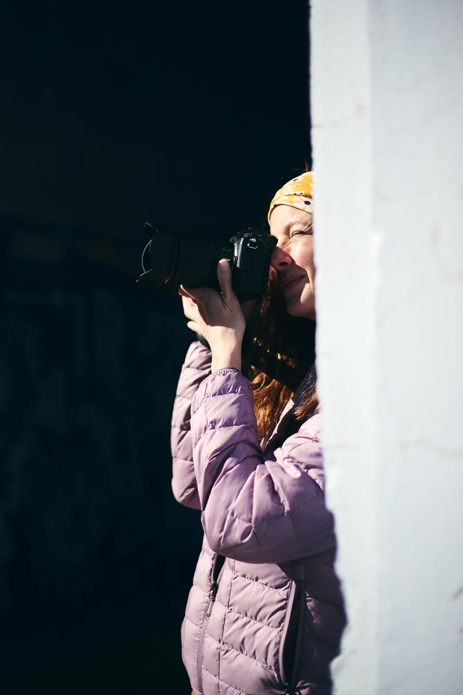 Young woman in pink jacket and yellow hat photographing with DSLR camera outdoors against white wall.