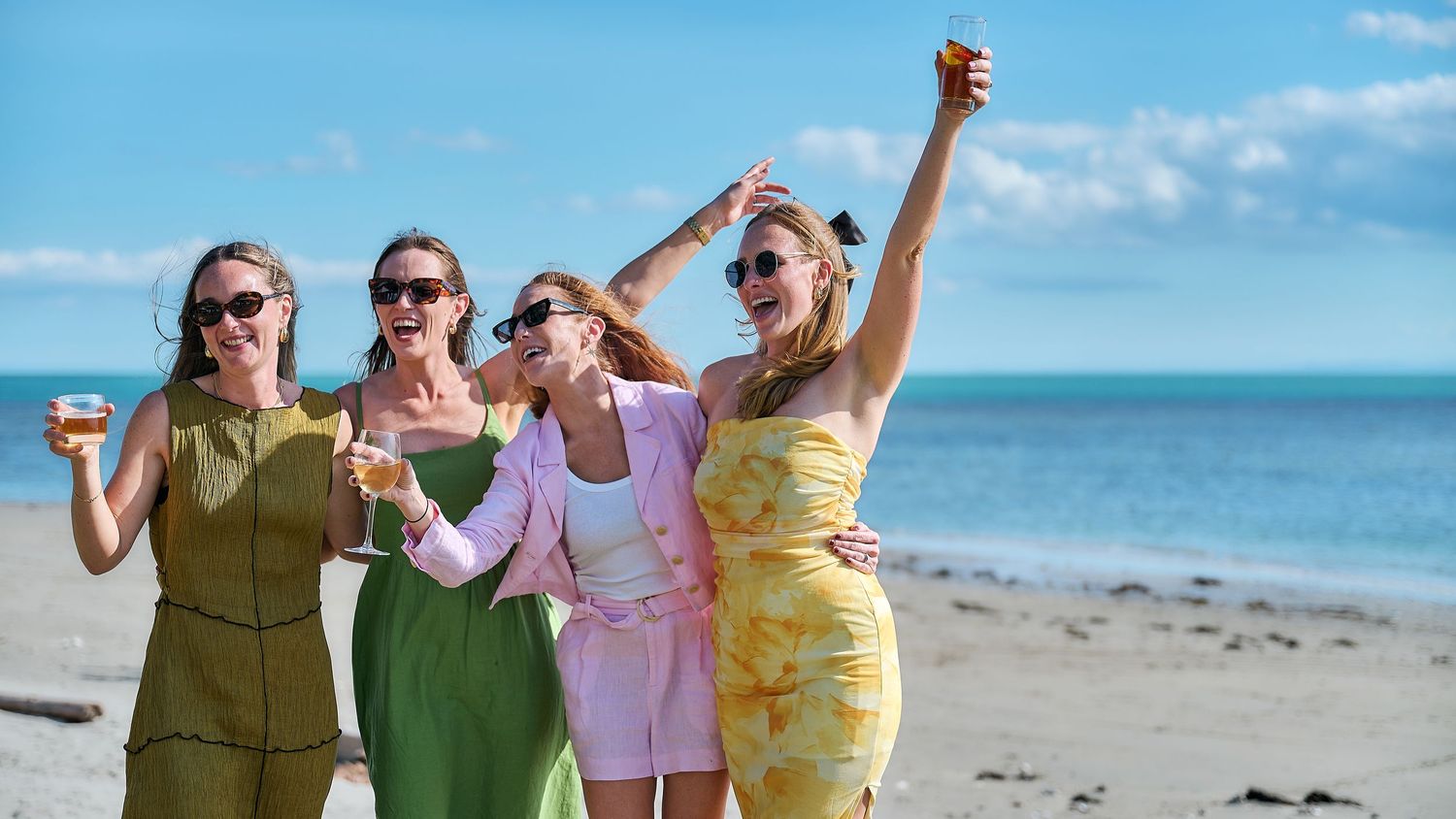 Four stylish women celebrating on a beach, holding drinks and dancing in colorful summer outfits.