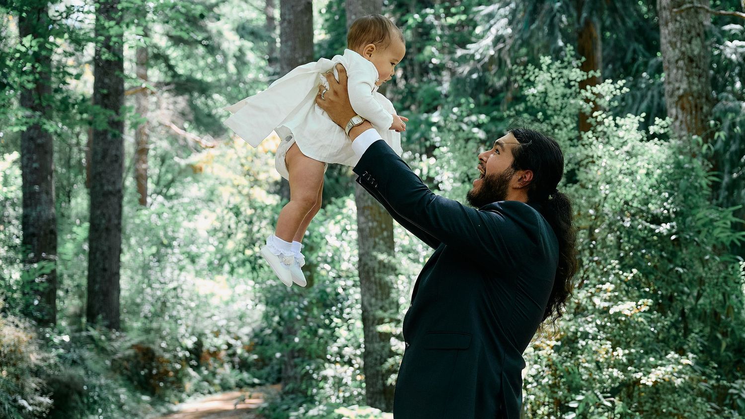 Bearded father in a dark suit lifting a baby in white clothing in a sunlit forest.