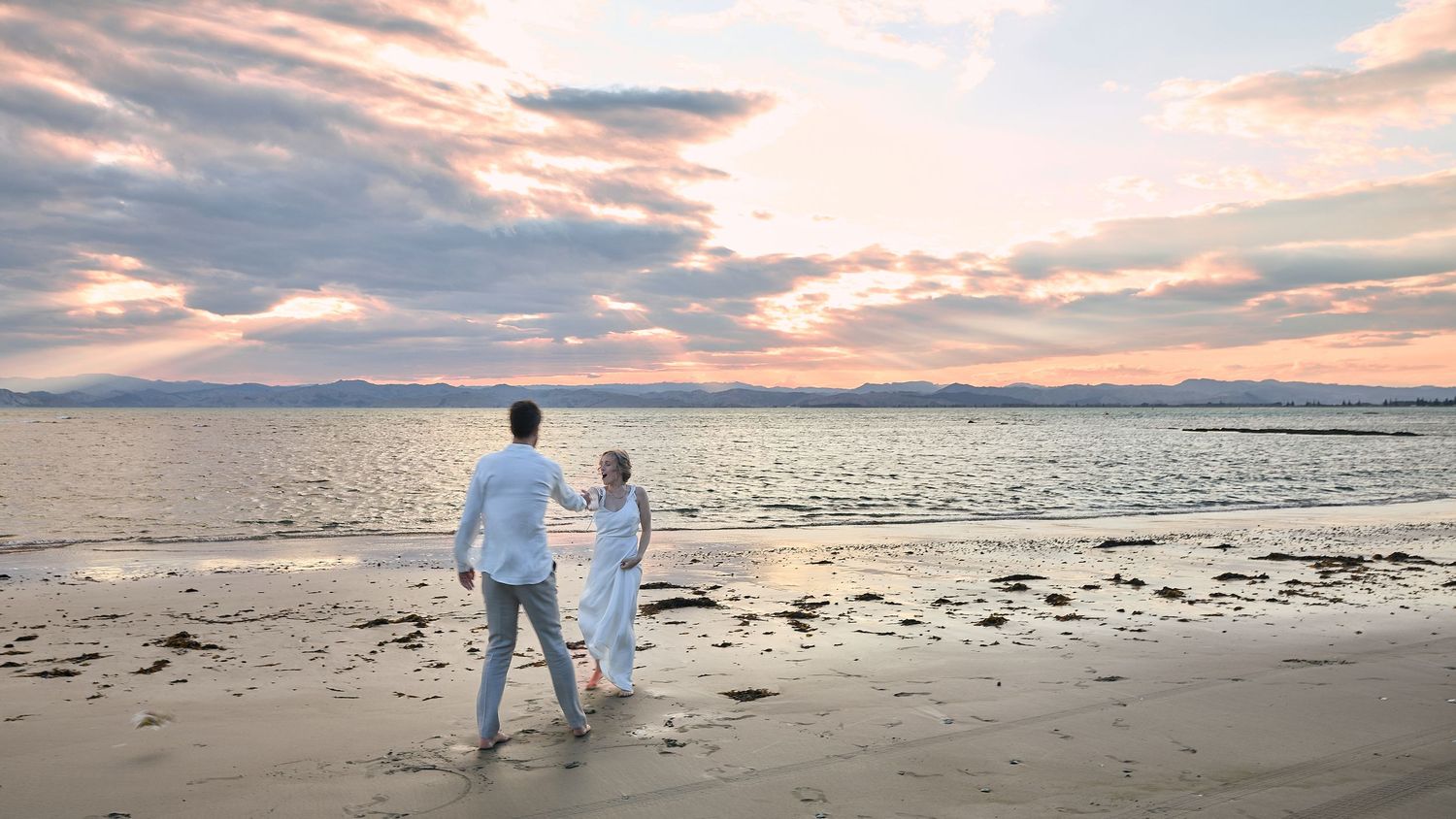 Couple walks hand in hand along a serene beach at sunset with mountains in the distance.