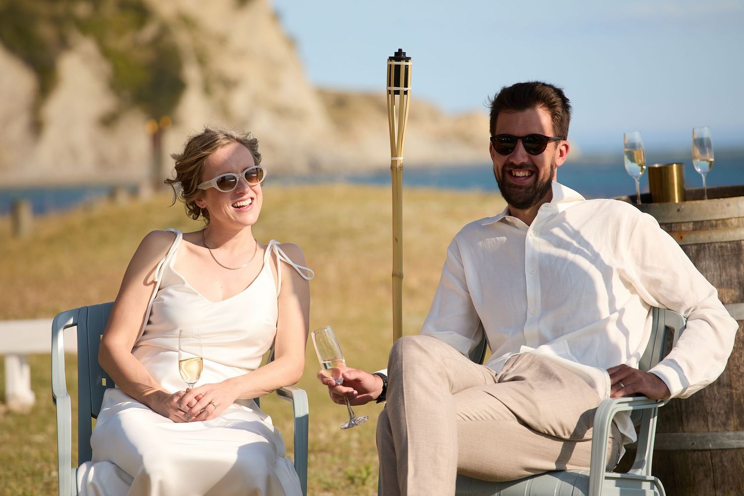 Bride and groom in sunglasses relax in chairs with champagne at outdoor coastal wedding reception.