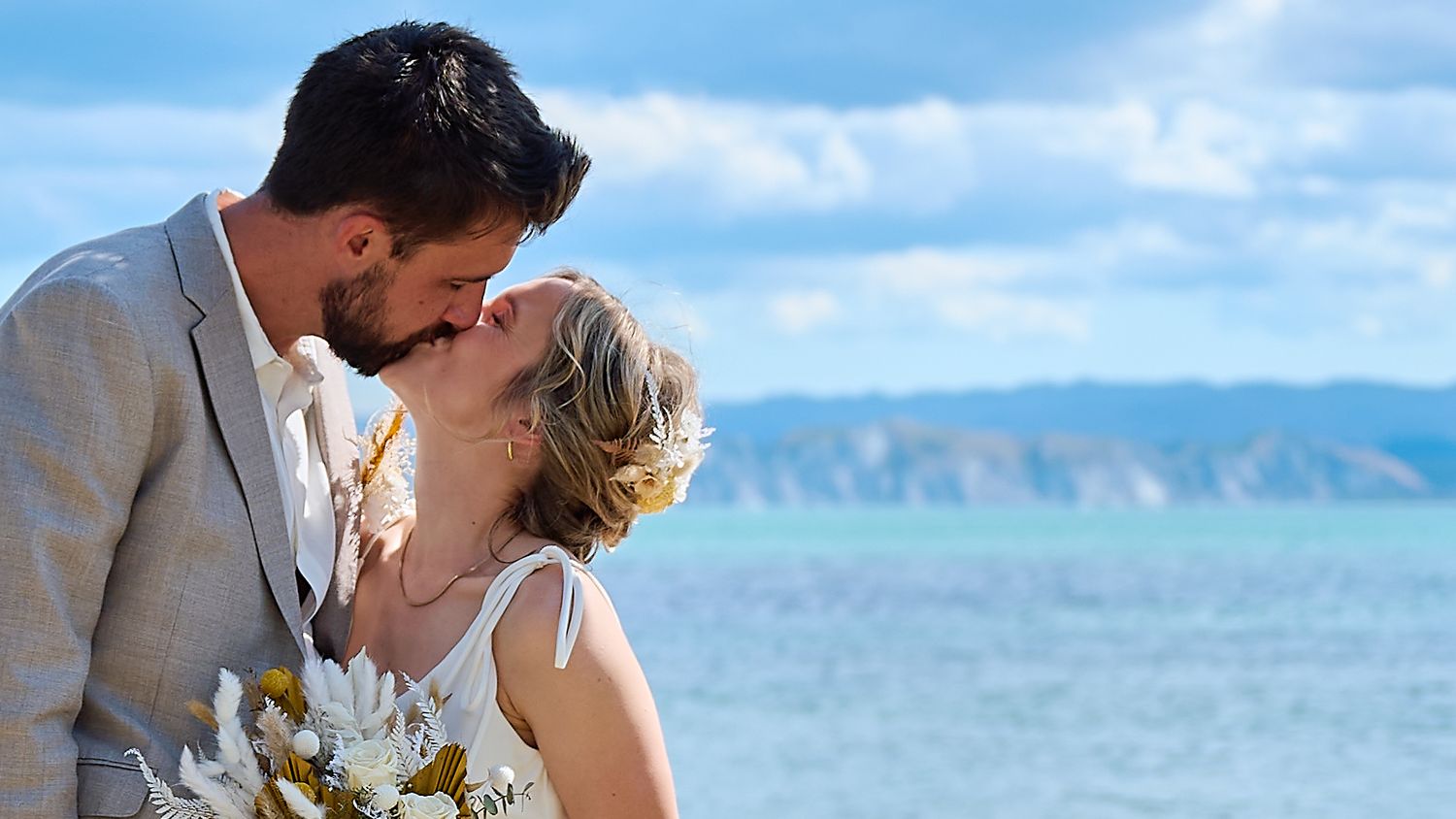 Bride and groom sharing a romantic kiss on the beach with turquoise ocean and blue sky in the background.