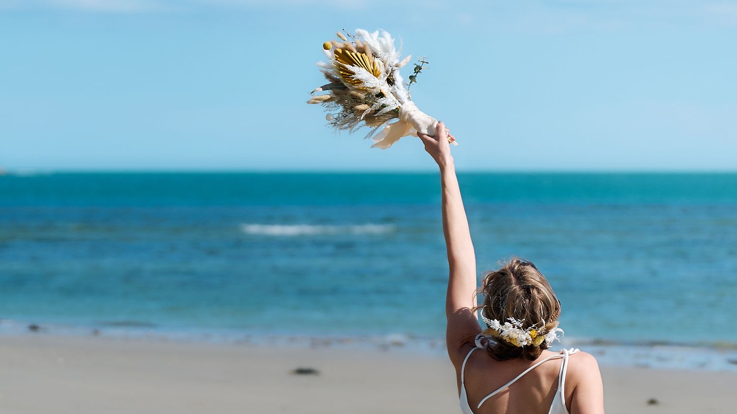 Bride raises dried flower bouquet toward blue ocean sky at beach wedding.