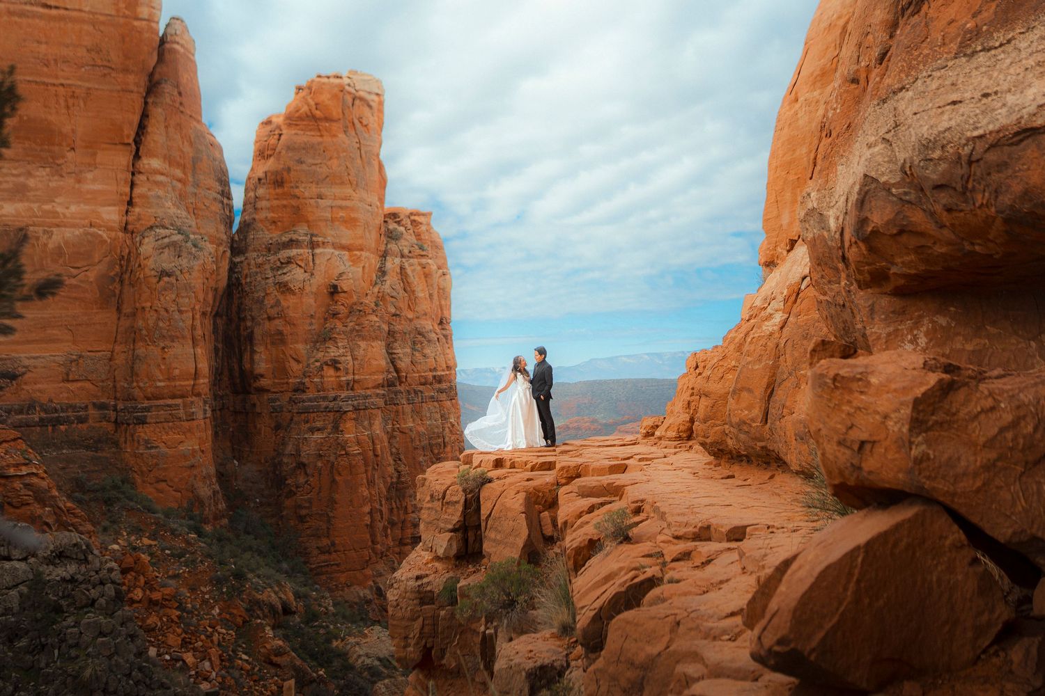 Bride and groom standing on a red rock ledge with a sweeping canyon view in Sedona.