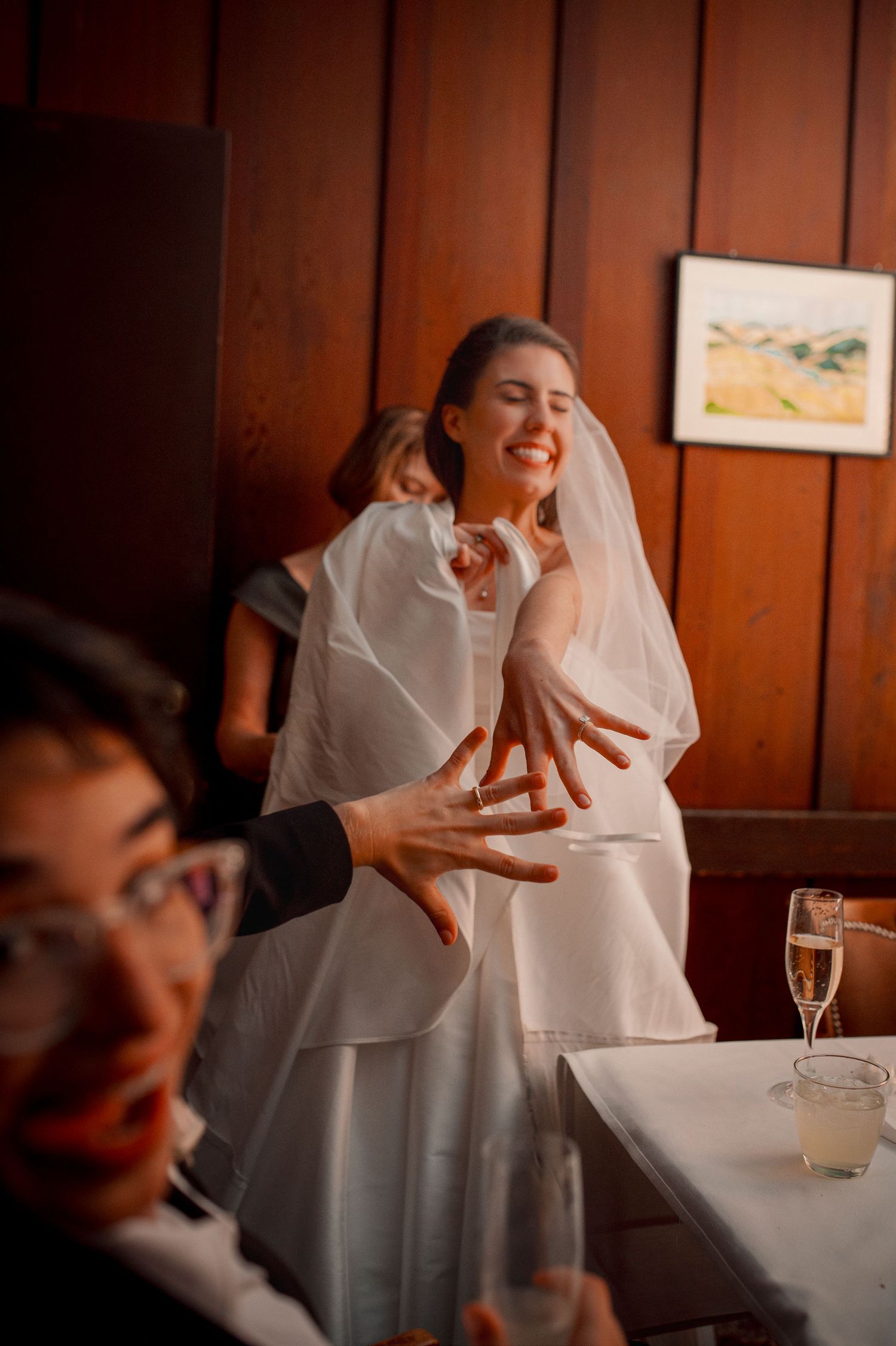 Joyful bride in a white veil laughing and gesturing with her hands at a wedding reception.