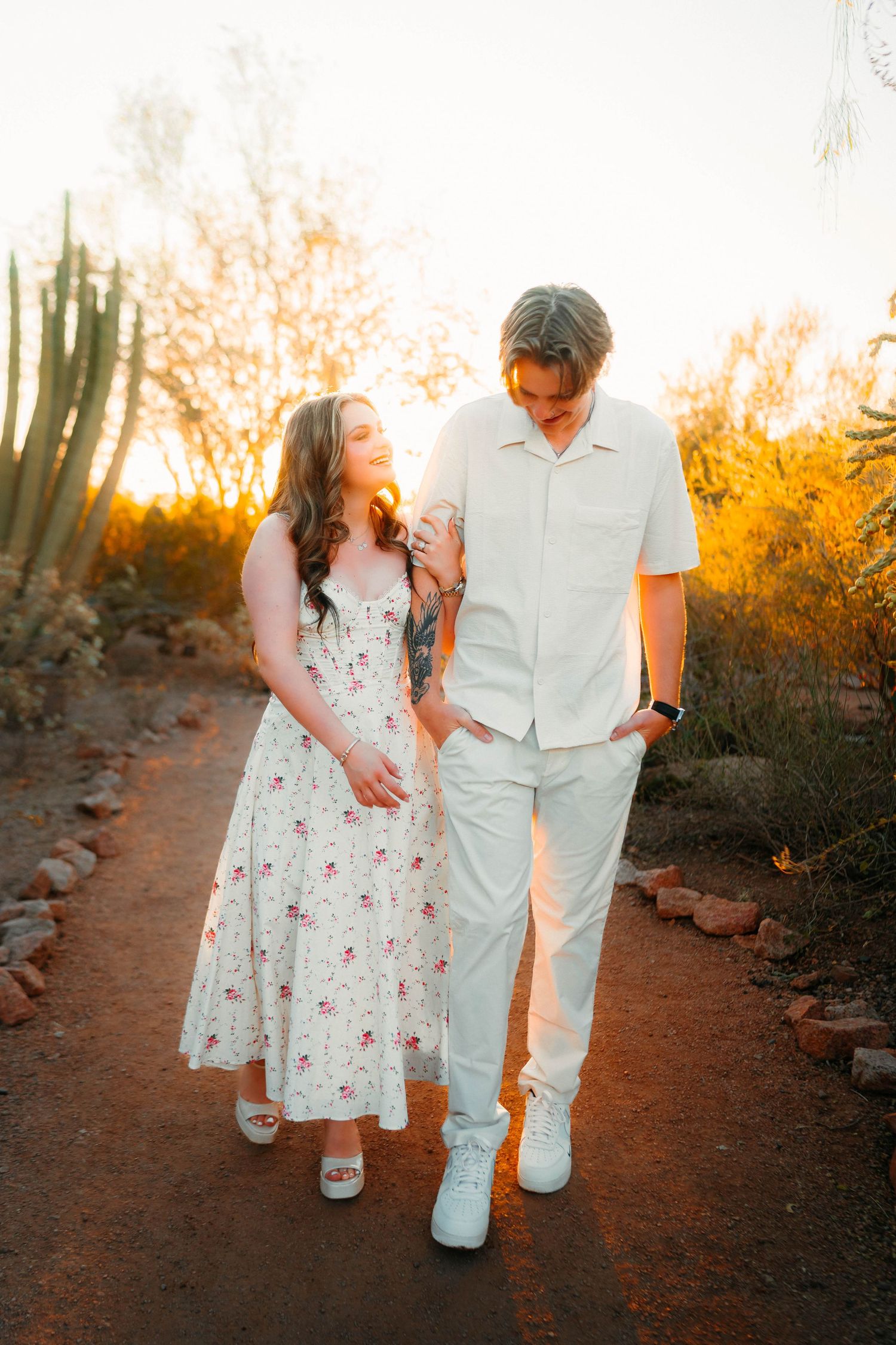 Couple walking hand in hand at sunset in a desert garden, woman in floral dress, man in white outfit.
