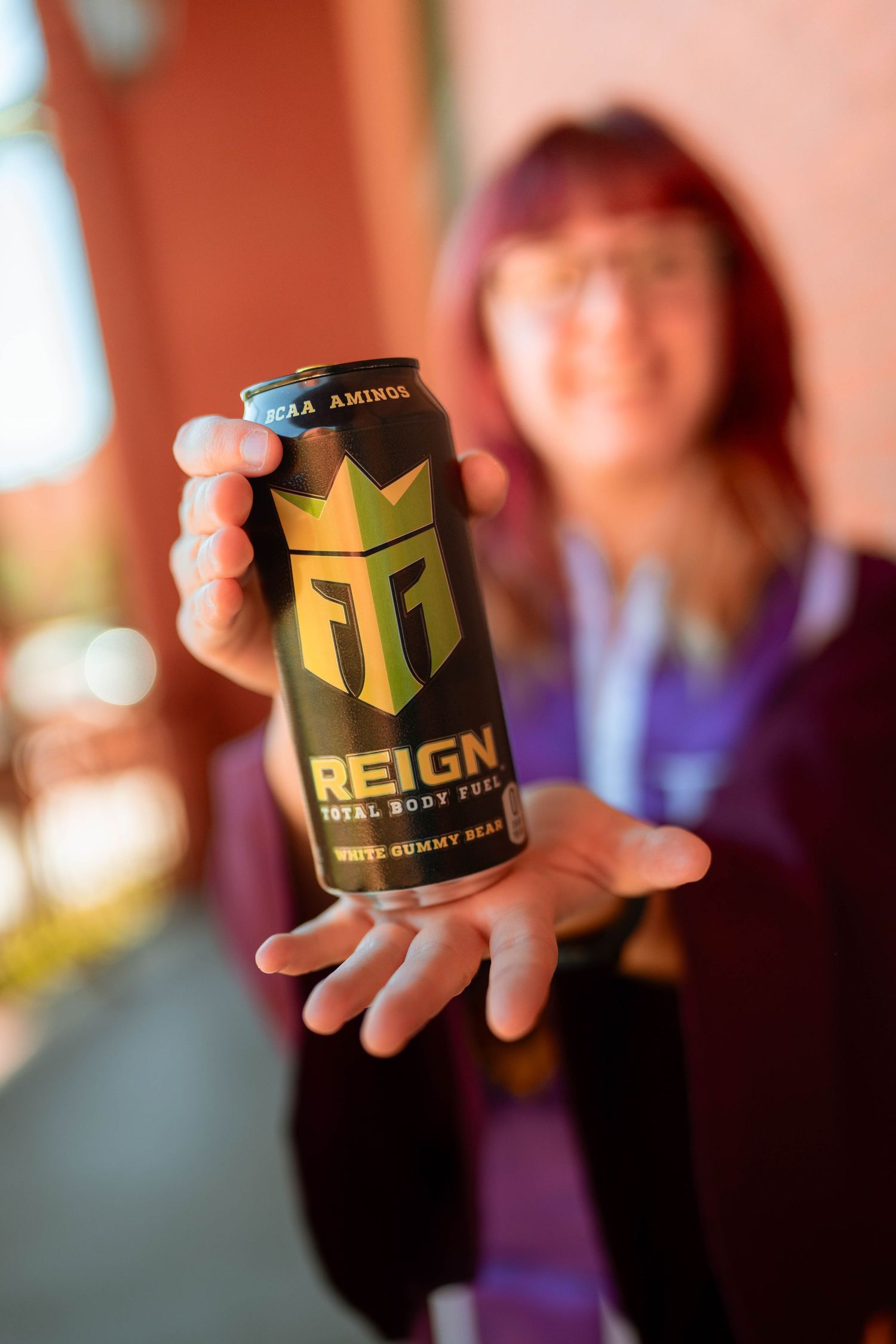 Woman with red hair holding a can of Reign Total Body Fuel White Gummy Bear energy drink toward the camera.