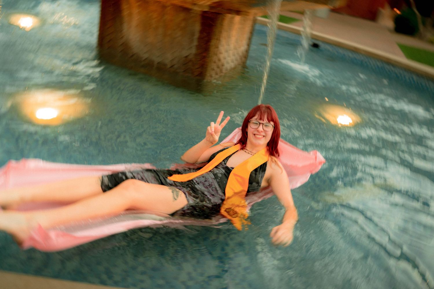 Young woman with red hair relaxing on a pink float in an indoor pool giving a peace sign.