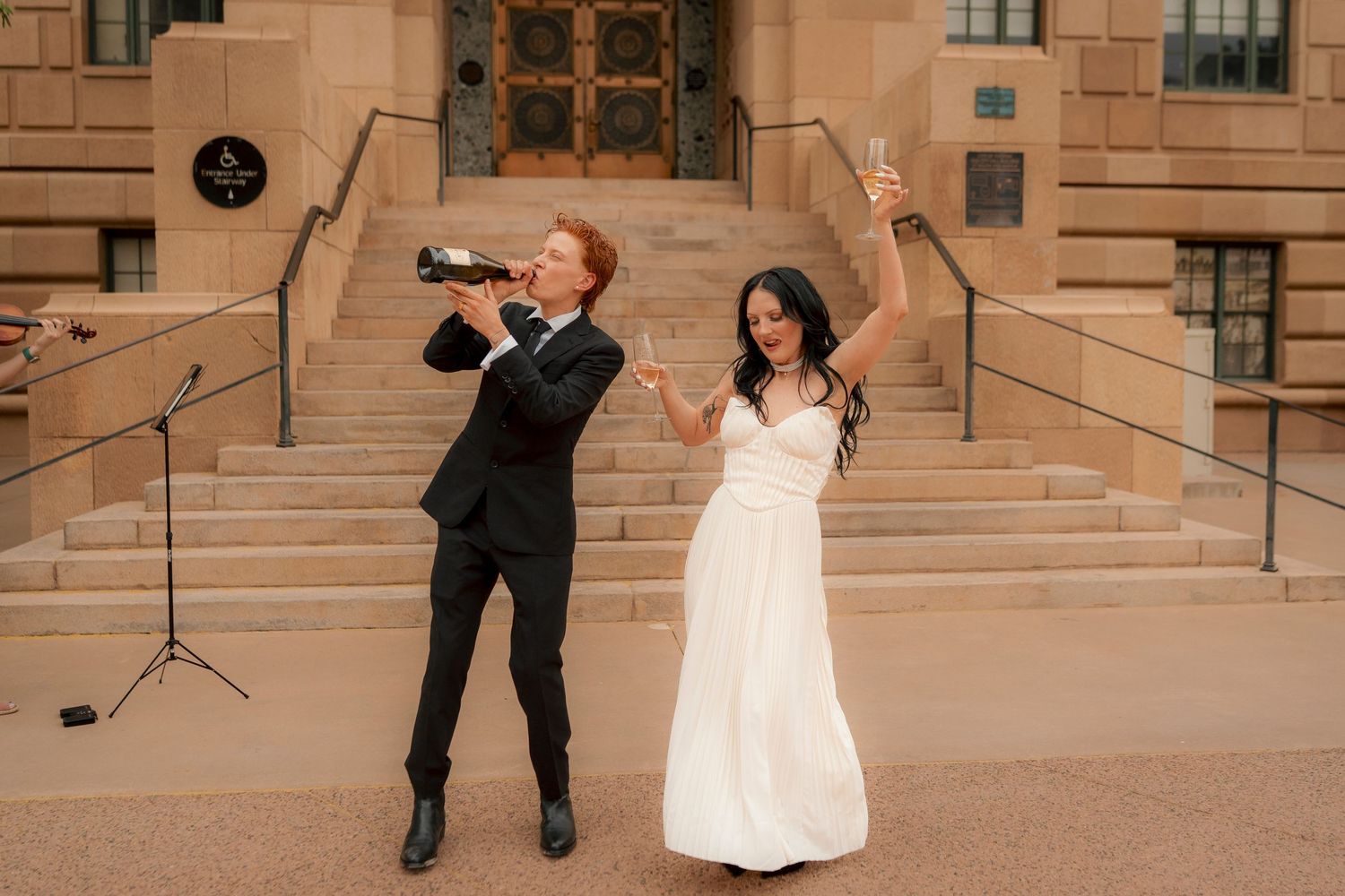 Joyful newlyweds celebrating with champagne bottle outside grand stone building after ceremony.
