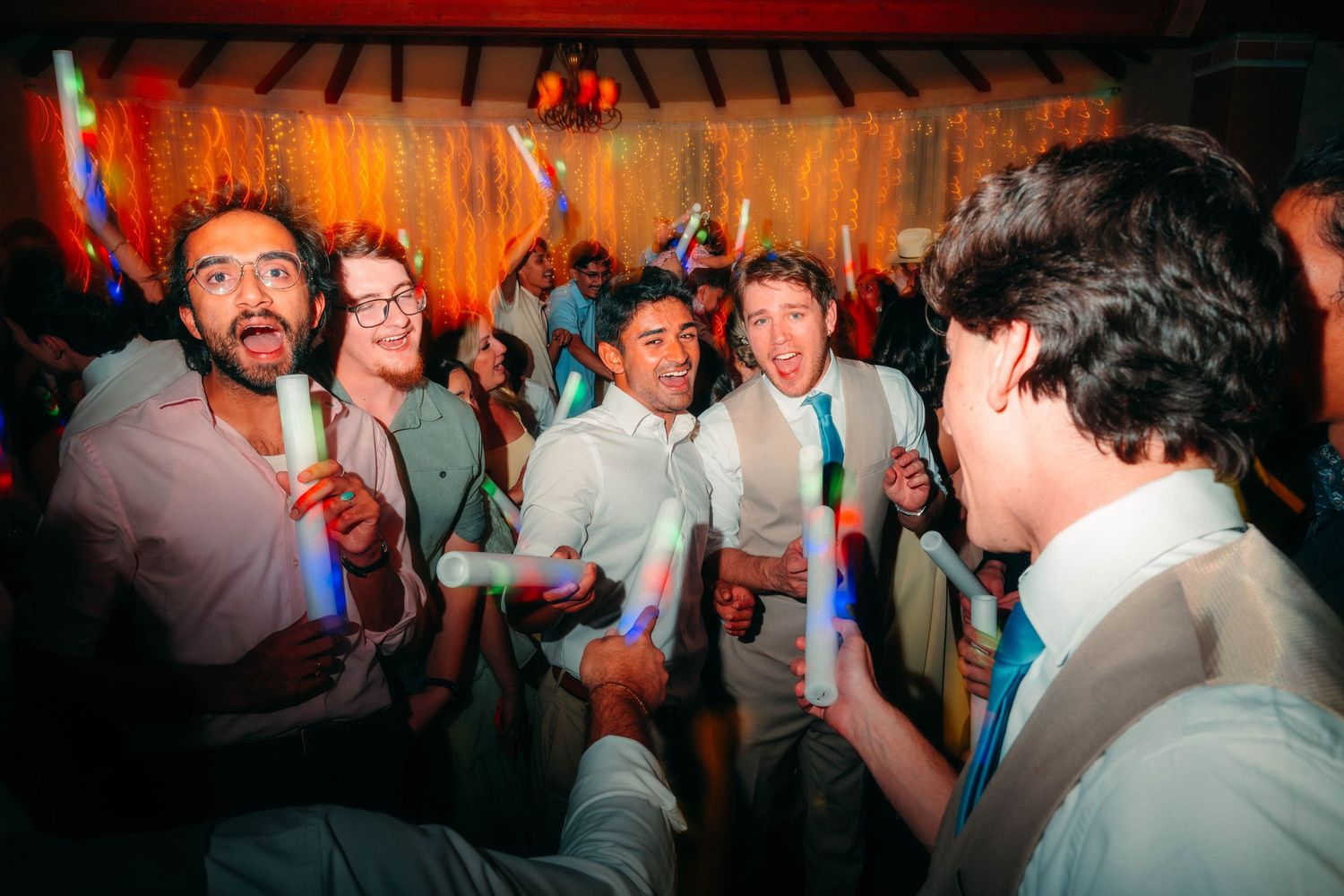 Joyful wedding reception guests dancing and celebrating with colorful glow sticks at a lively indoor party.