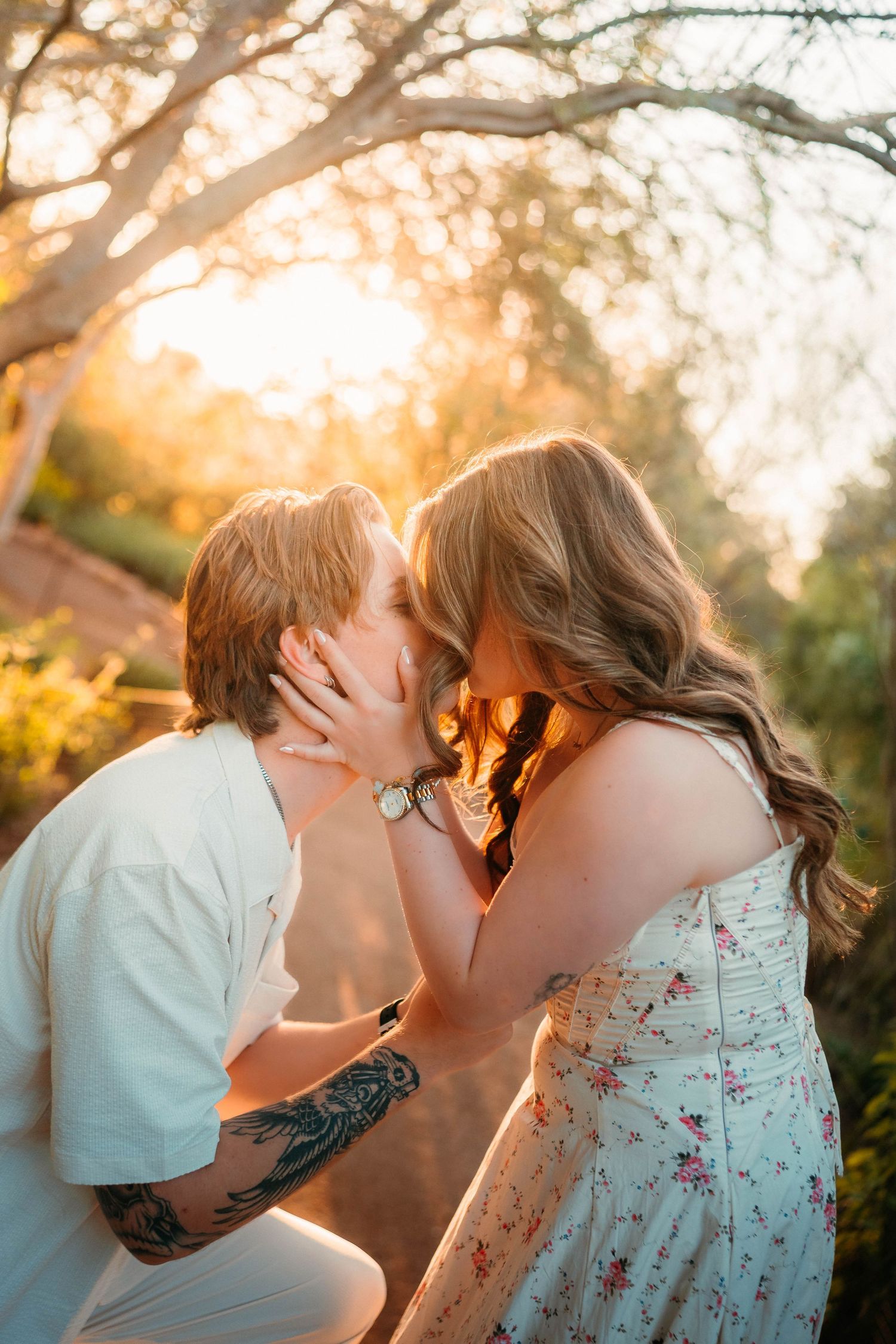Couple sharing a romantic kiss at golden hour with warm sunlight filtering through trees.