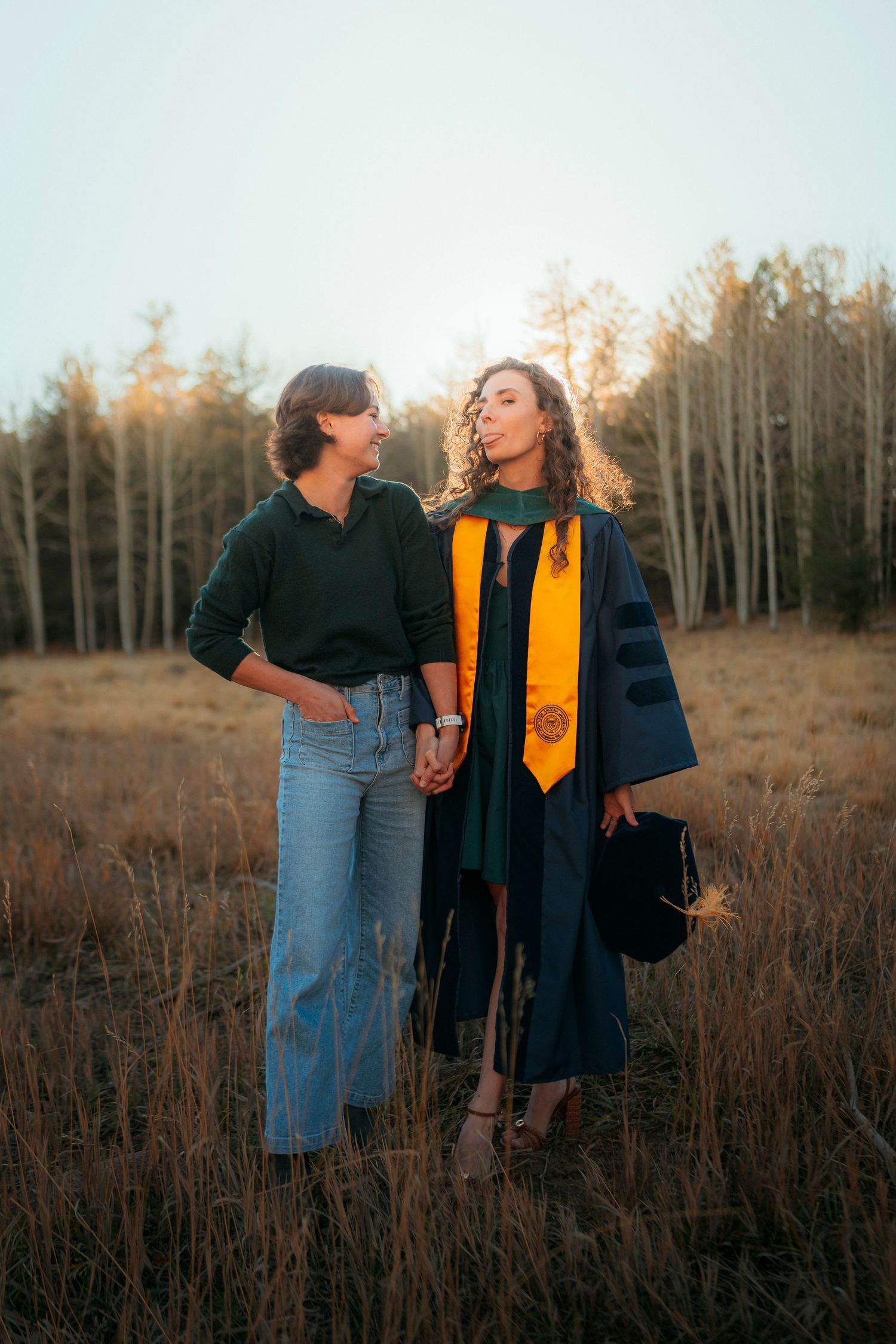 Graduate in navy and gold regalia holds hands with partner in sunlit autumn forest field.