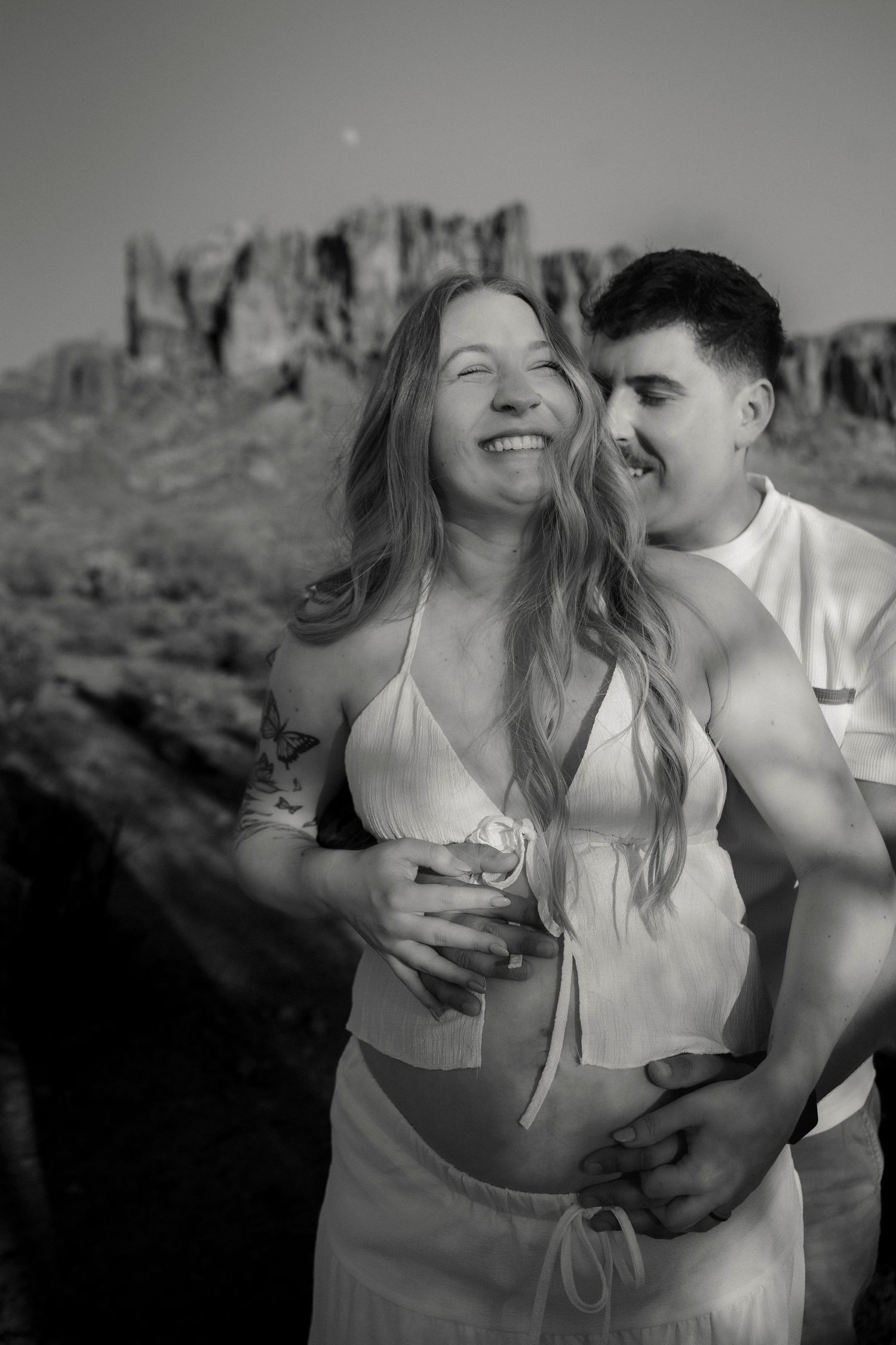 Black and white photo of smiling pregnant couple embracing outdoors with rocky desert mountains behind.