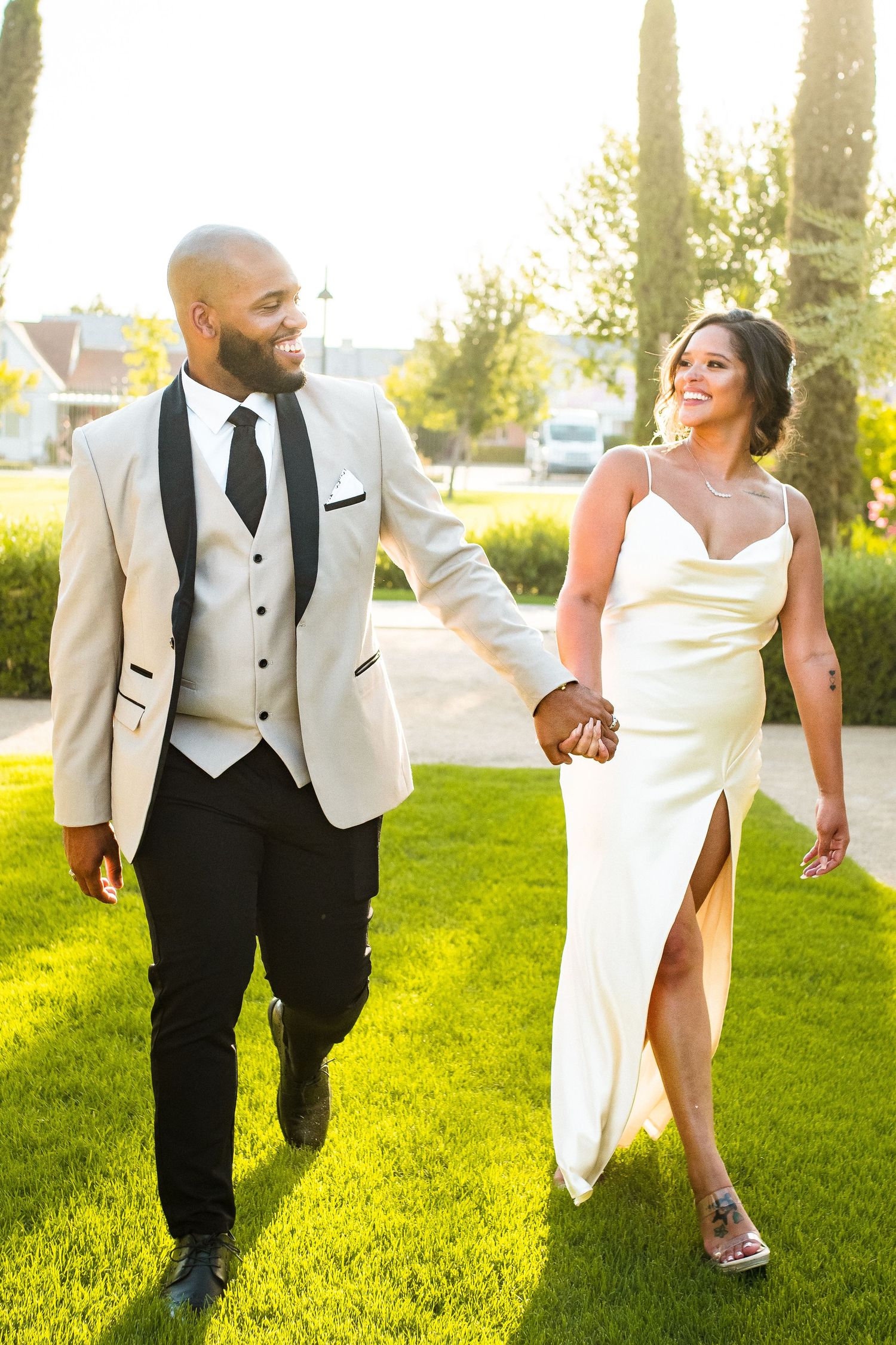 Newlywed couple holding hands and smiling at each other while walking on green grass in golden sunlight.