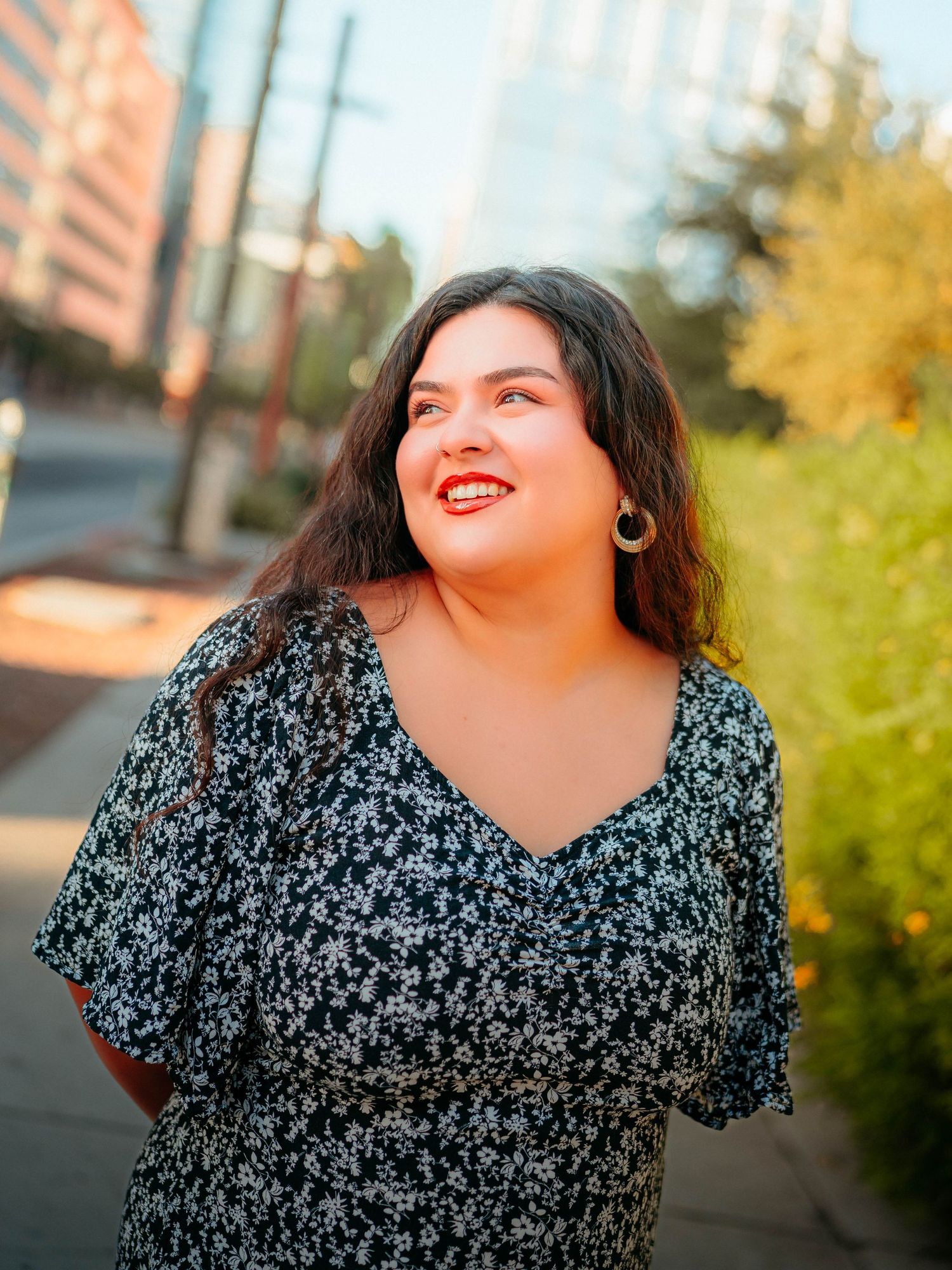 Smiling brunette woman in floral dress standing on urban sidewalk at golden hour.