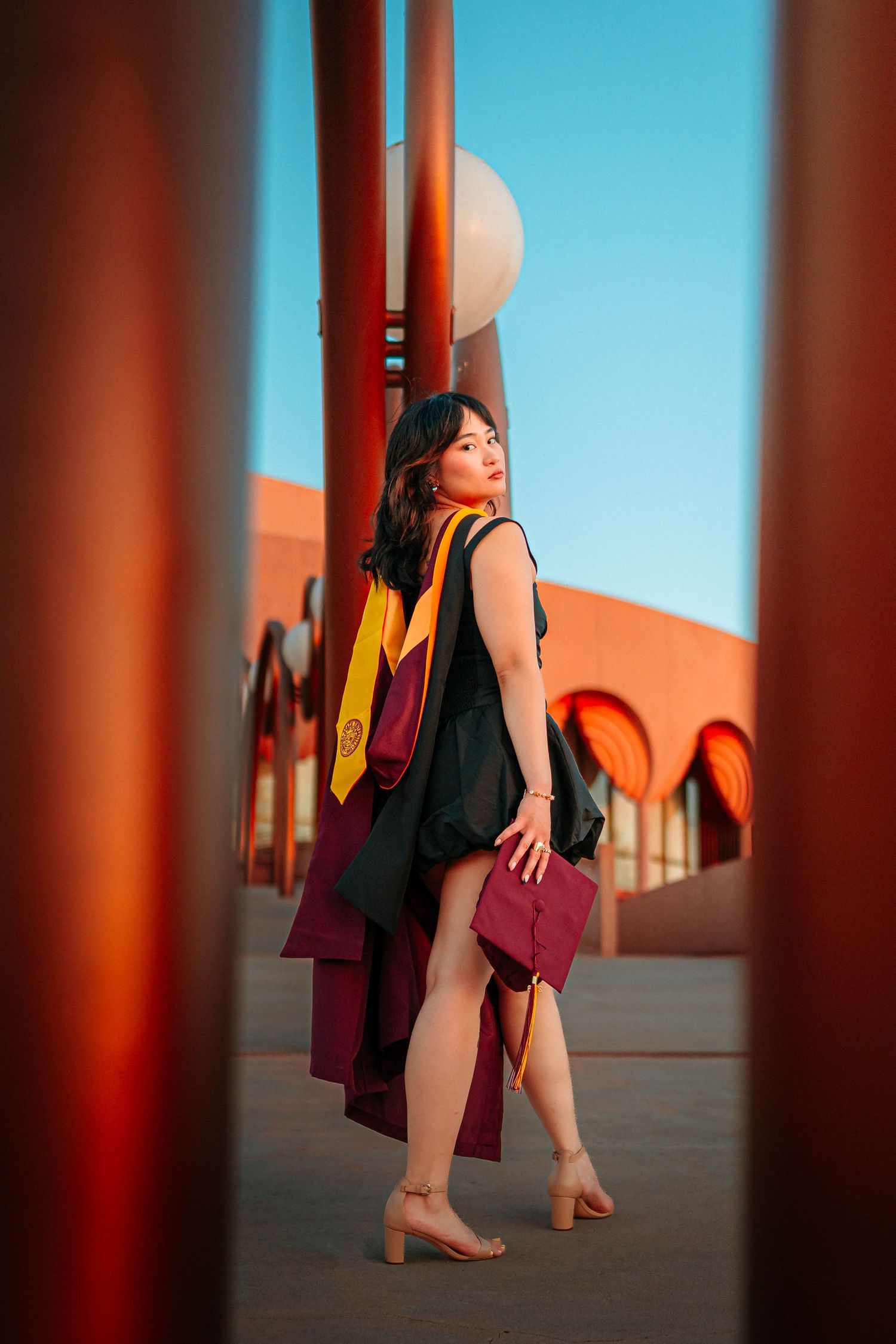 Young woman in ASU master's graduation gown with gold sash holds maroon cap outdoors near red columns under blue sky.