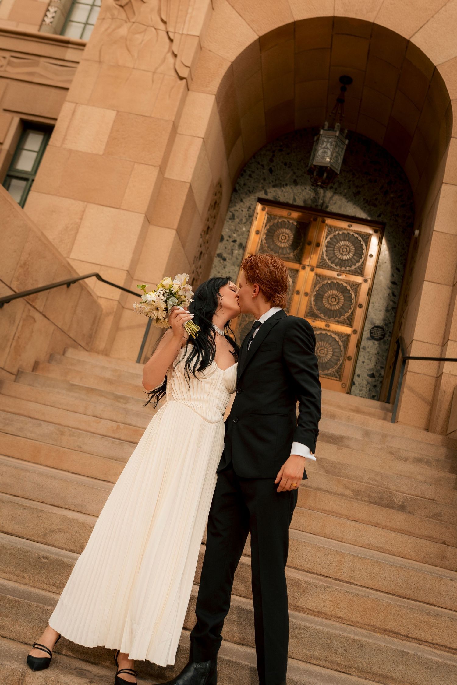 Bride and groom kissing on steps of ornate building, bride in white pleated dress holding bouquet, groom in black tuxedo.