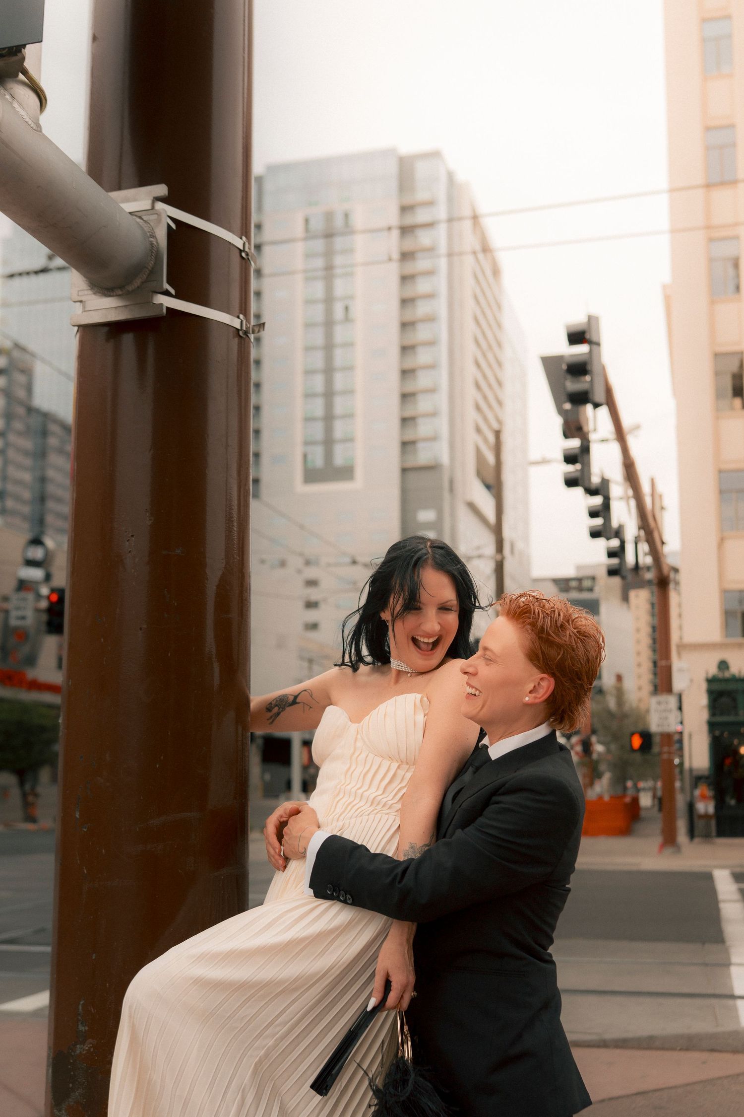 Laughing bride in white pleated dress sitting on street pole, groom in black suit embracing her at urban city intersection.