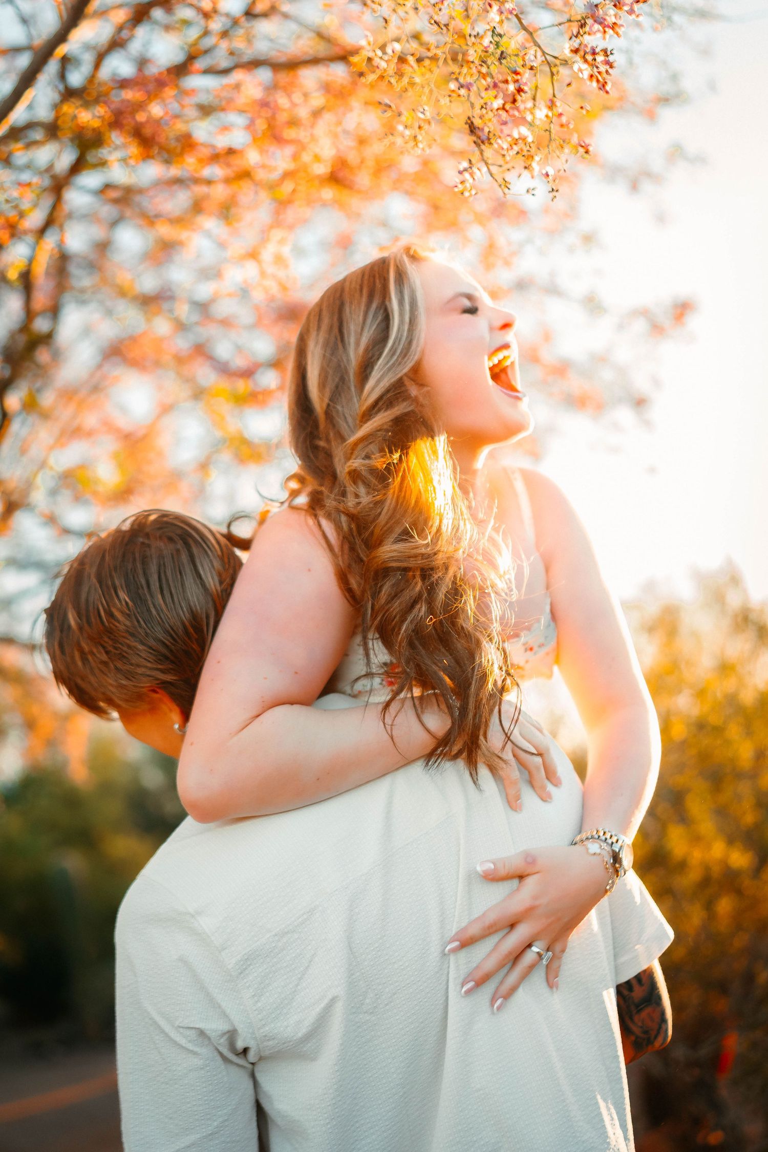 Couple laughing joyfully outdoors in autumn sunlight, woman in white dress being lifted, golden fall foliage background.