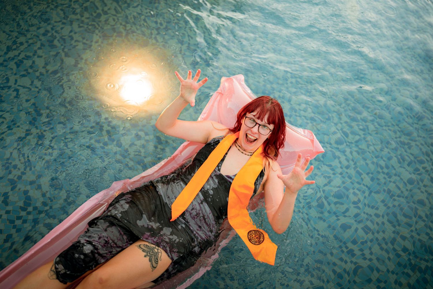 Woman in camo outfit floating on pink pool raft in blue pool, wearing orange sash and glasses.