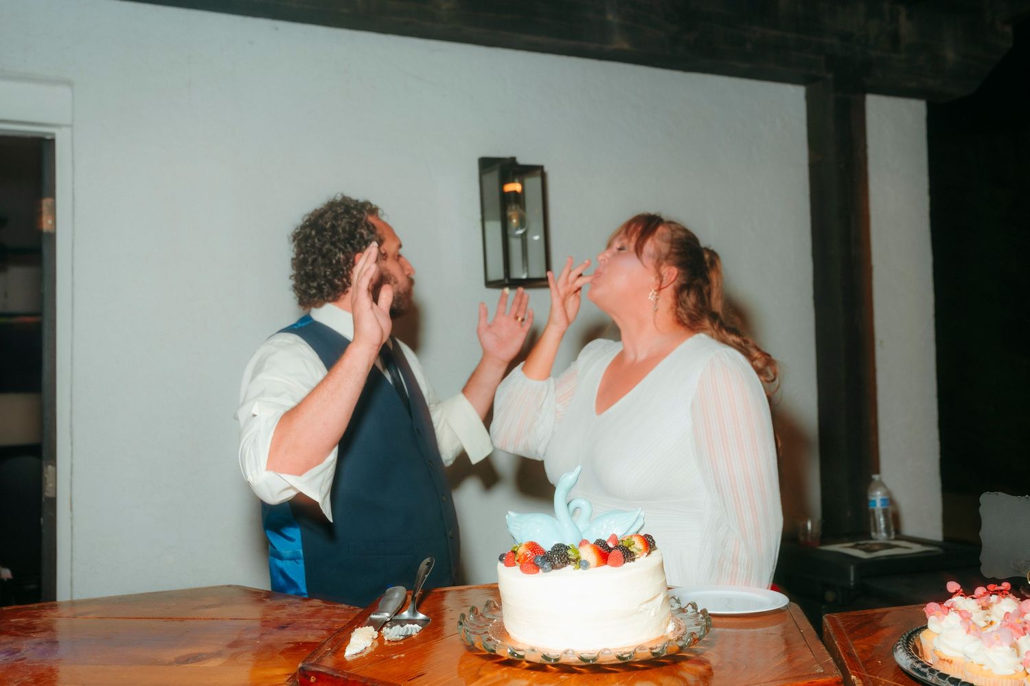 Bride and groom joyfully eating wedding cake together at rustic reception table.