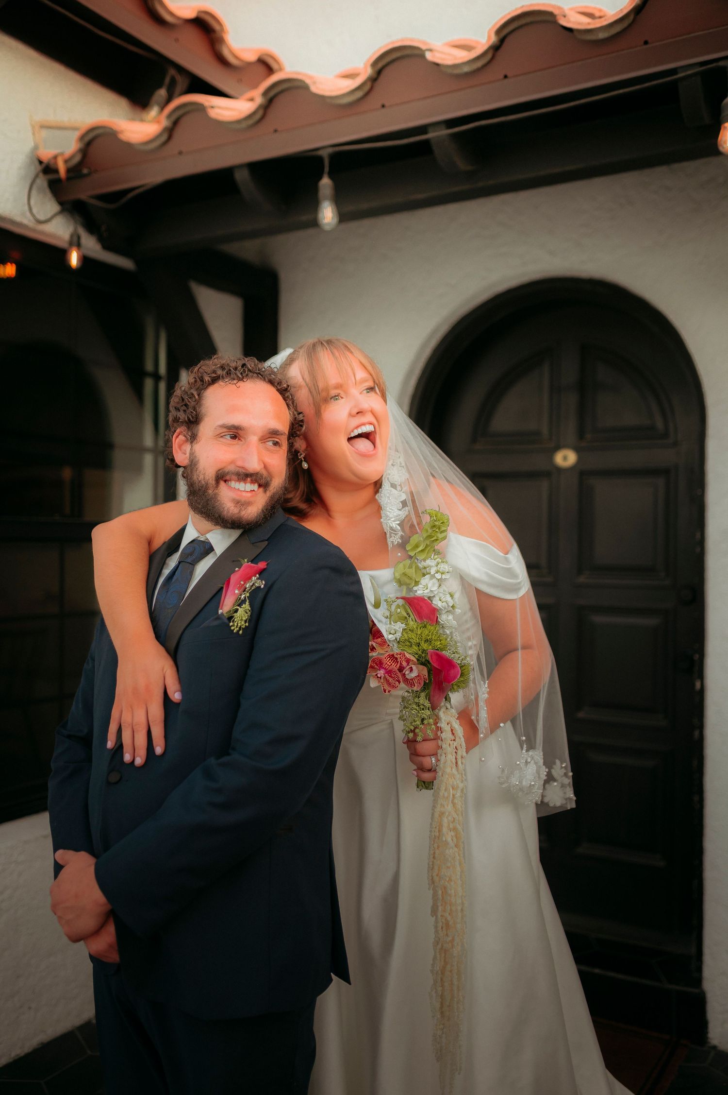 Joyful bride in white gown and veil holding floral bouquet embraces smiling groom in navy suit at wedding venue.