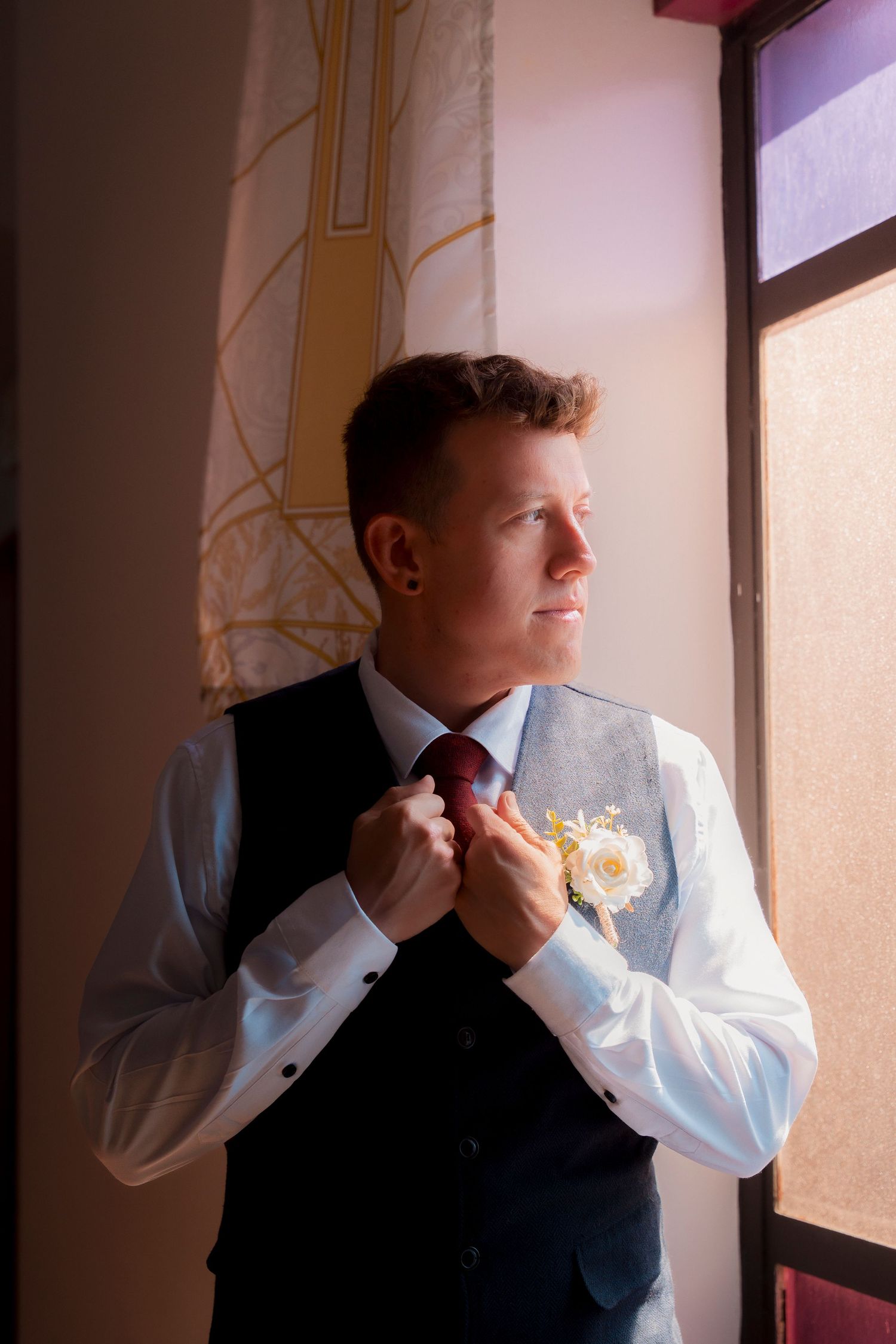 Groom adjusting his red tie wearing a vest with a white boutonniere by a window.