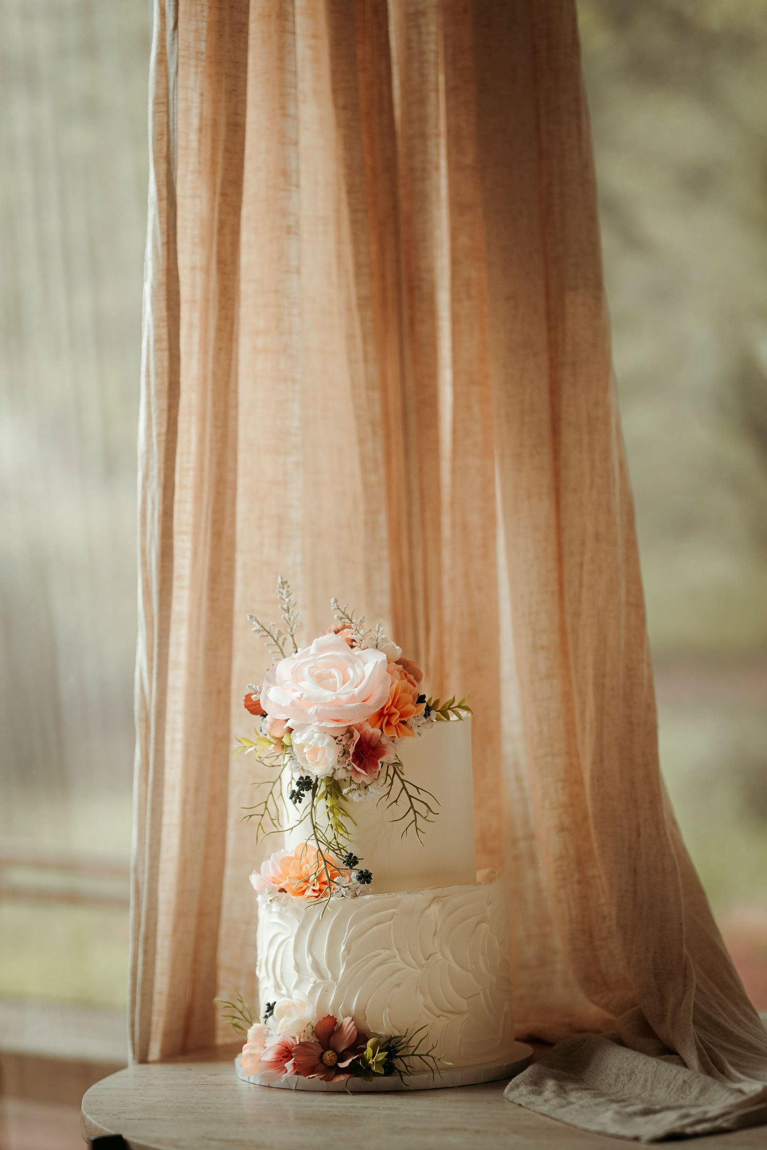 Elegant two-tier white wedding cake adorned with pink and coral flowers displayed before a soft peach curtain.