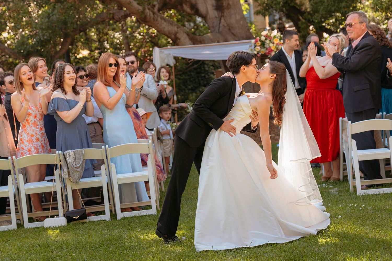 Bride and groom sharing a first kiss on the aisle as guests applaud at an outdoor wedding.