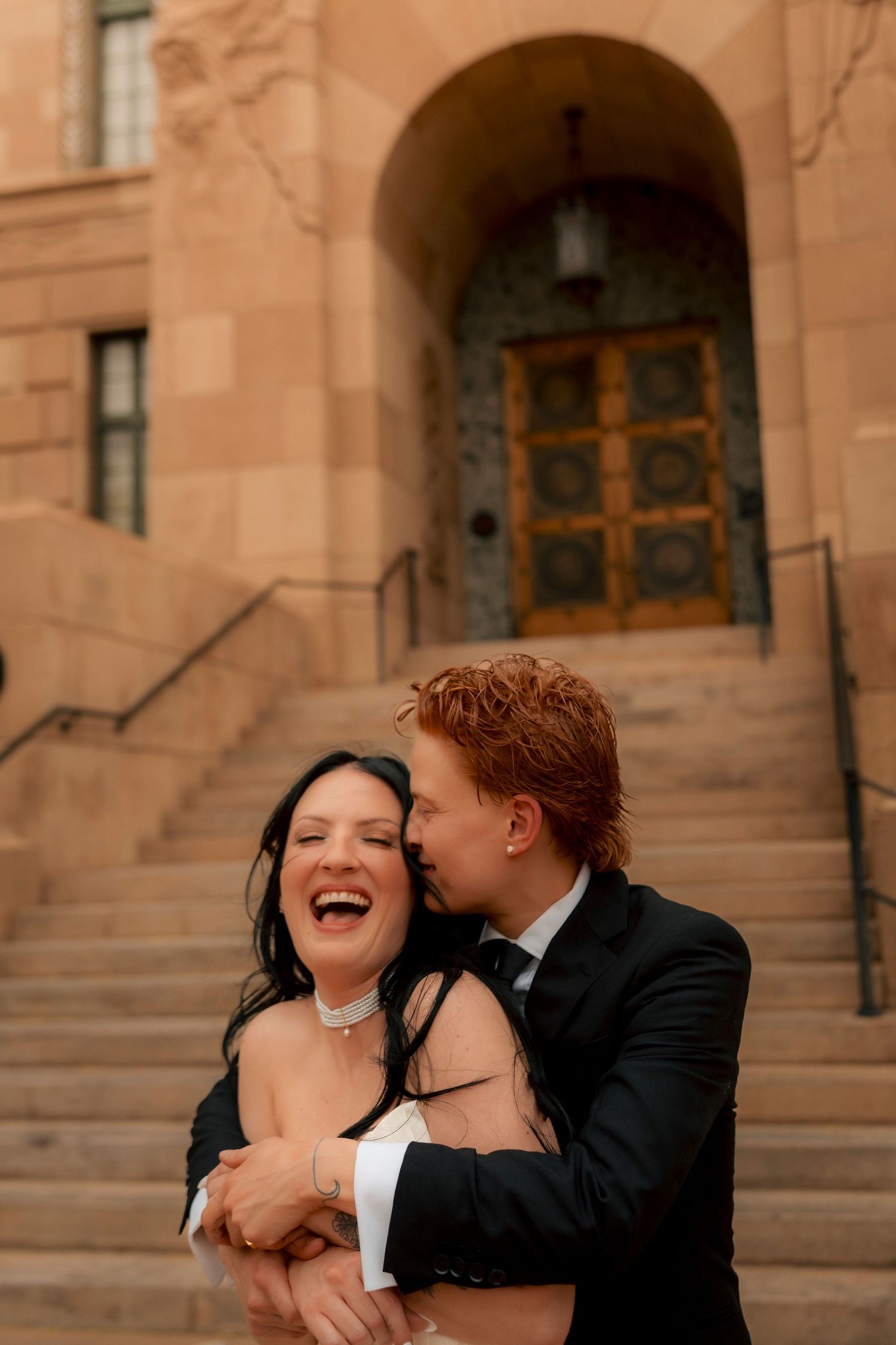 A joyful bride laughs as her red-haired groom kisses her cheek outside a grand stone building with arched entrance.