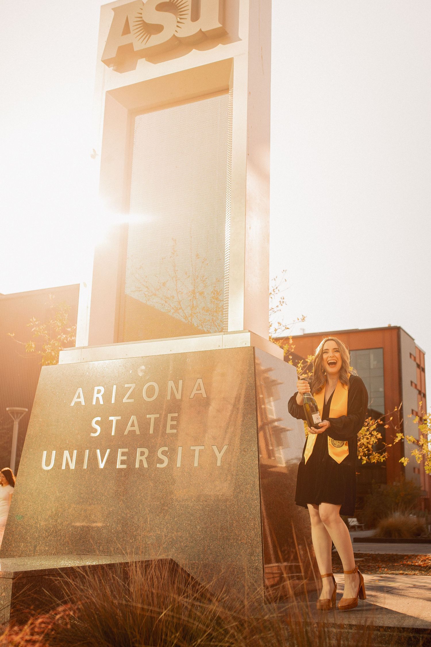 Proud graduate poses beside Arizona State University sign wearing black cap and gown at sunset.