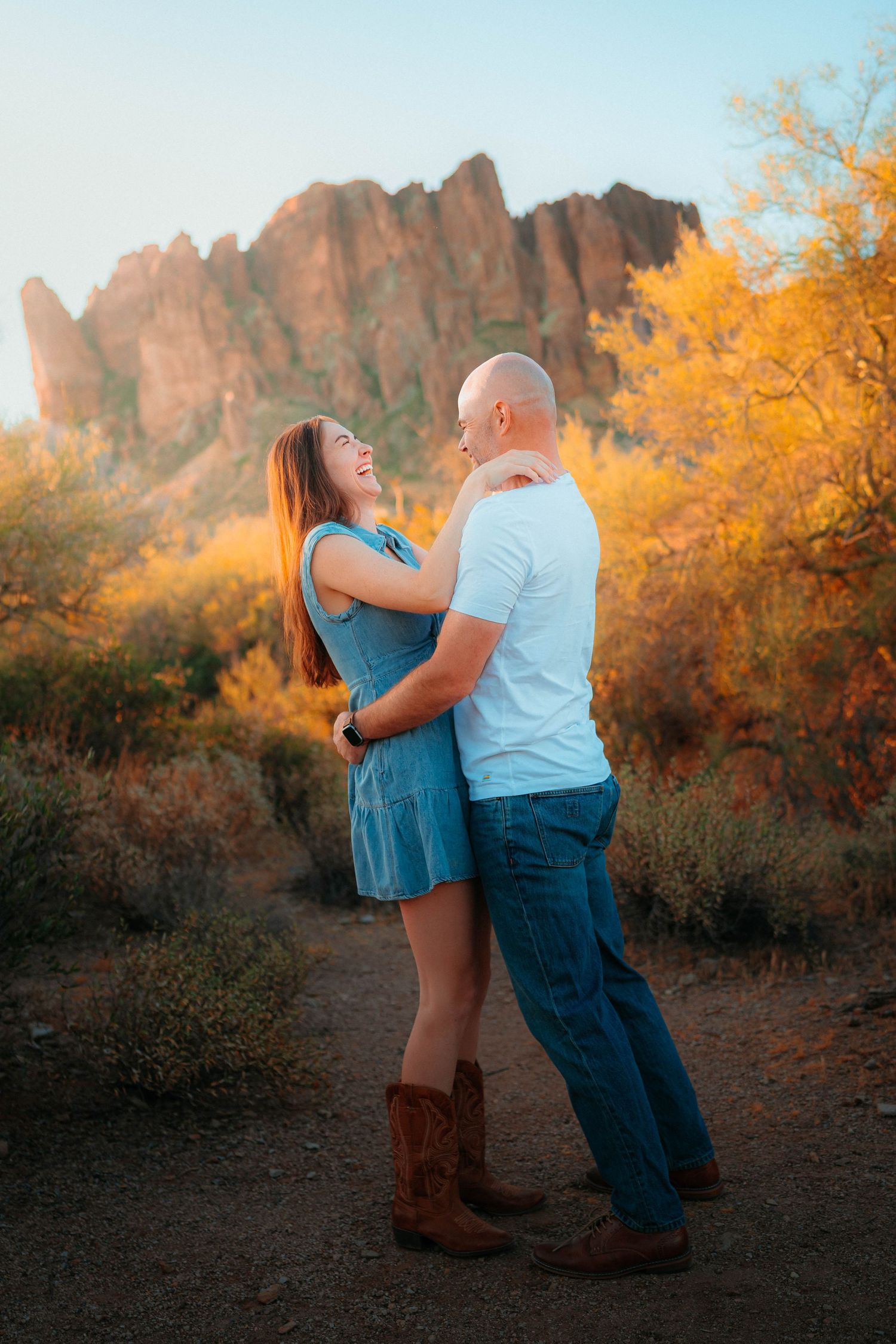 Laughing couple embracing on desert trail surrounded by golden foliage with Superstition Mountains at dusk.