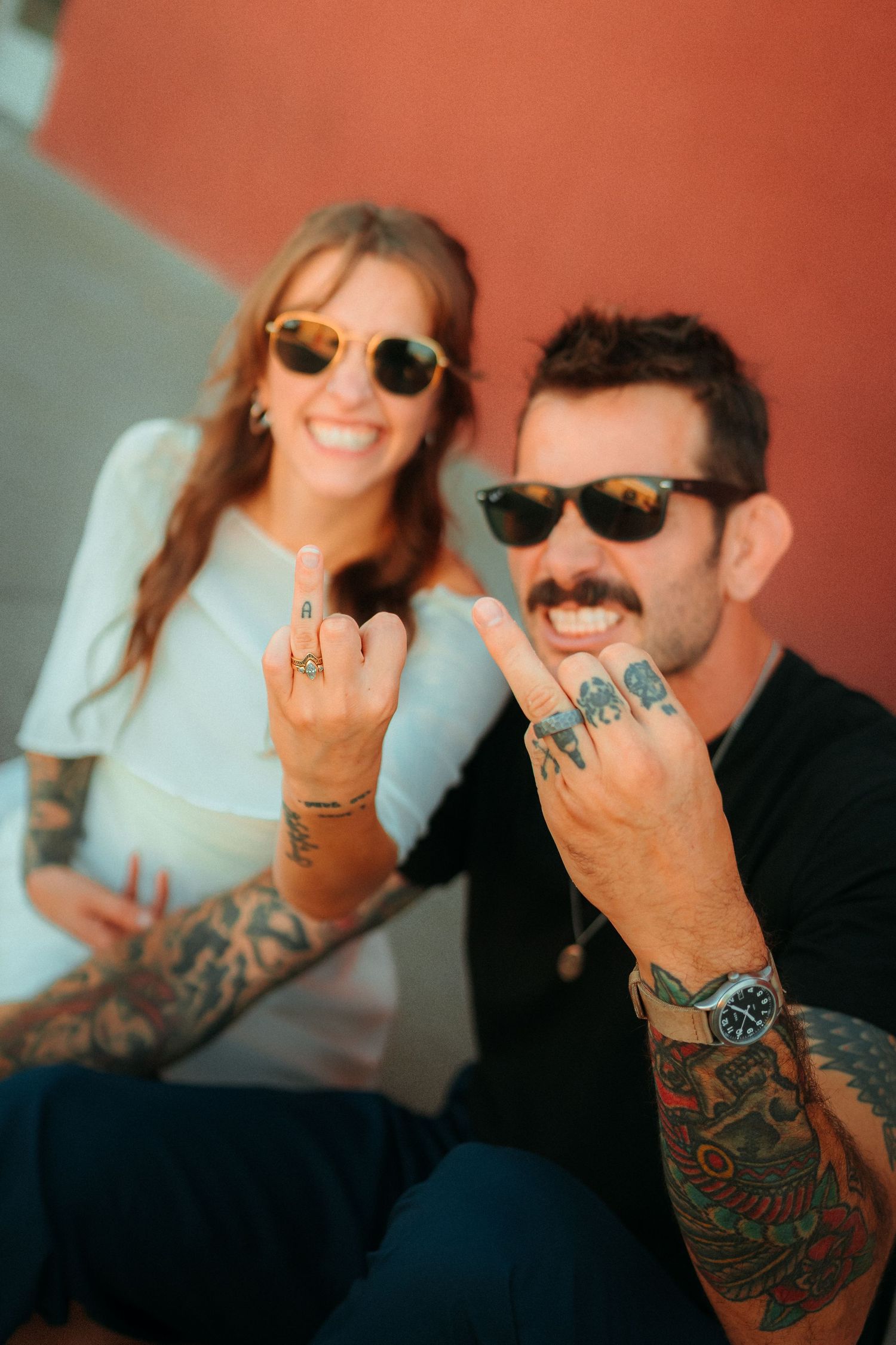 Tattooed couple in sunglasses smiling and showing off matching finger tattoos against a red wall.