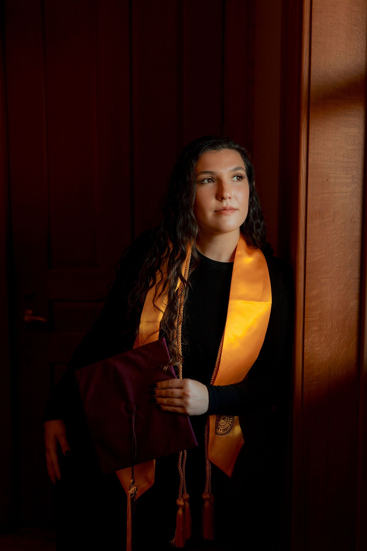Young woman holds maroon graduation cap wearing gold stole standing in dramatic doorway light.