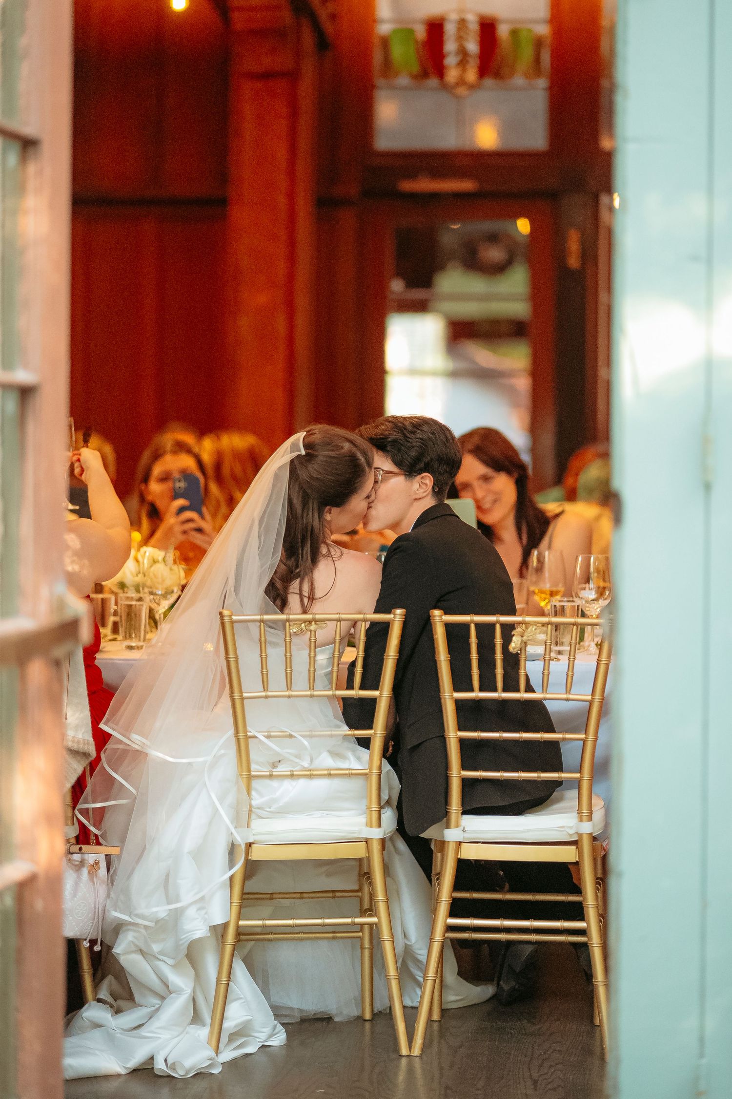 Bride and groom share a kiss at their wedding reception dinner table surrounded by guests.