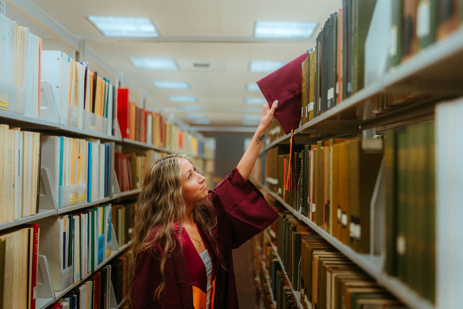 Graduate in cap and gown reaching for a book on a library shelf.