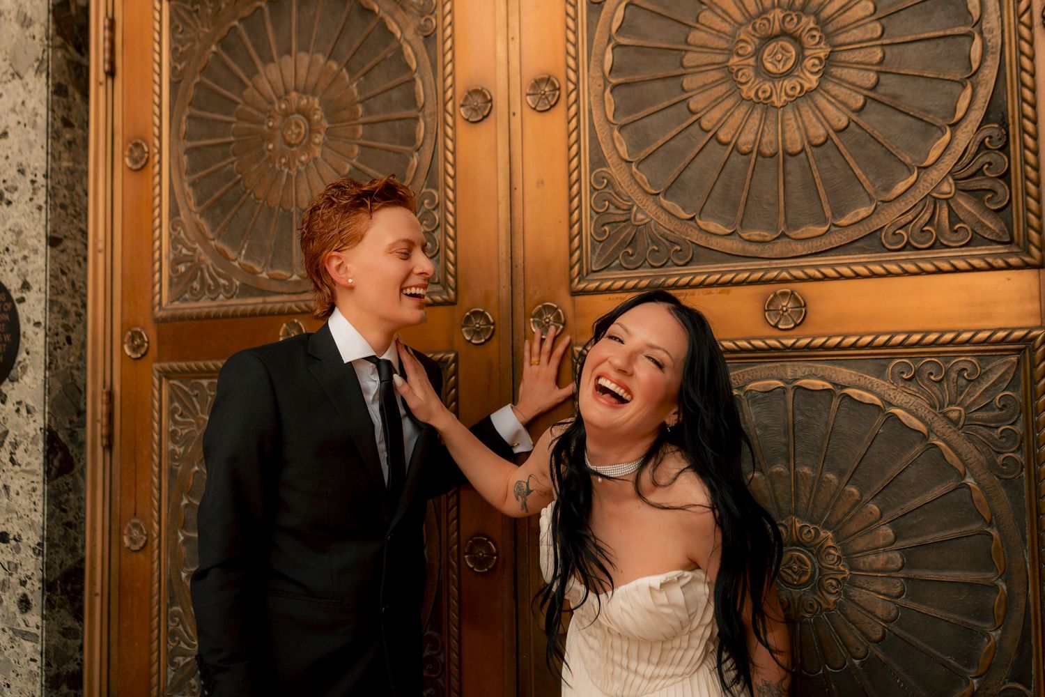 A laughing bride and groom pose playfully in front of ornate golden doors in an elegant venue.