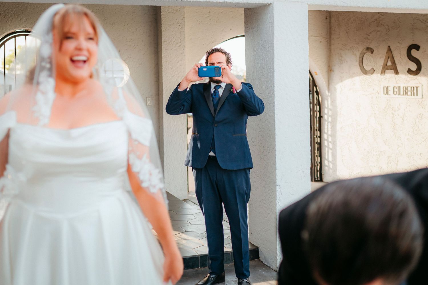 A man in a navy suit photographs a bride in a white gown and veil outside a wedding venue.