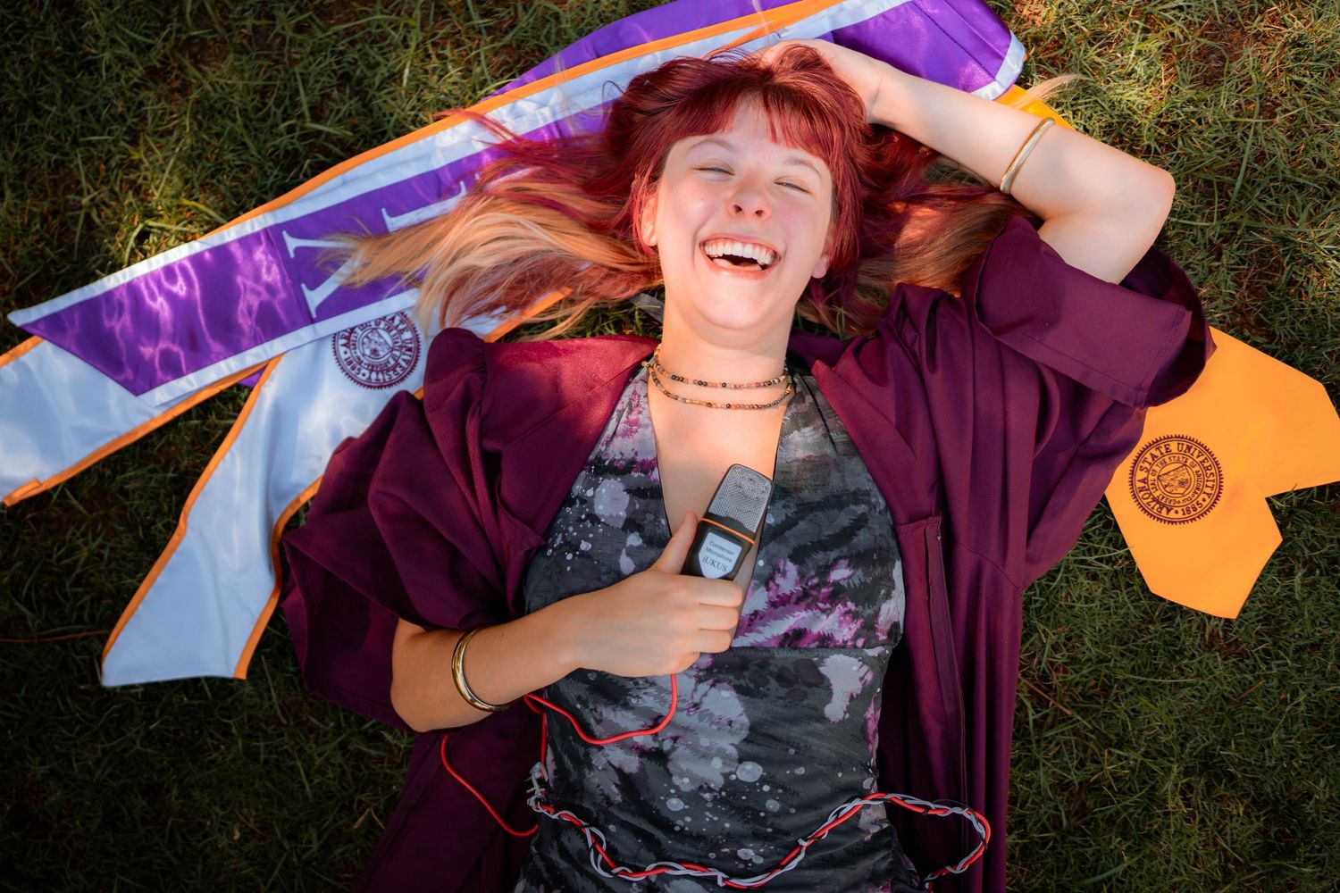 Red-haired woman laughing while lying on grass in maroon graduation gown with purple and blue academic sashes.