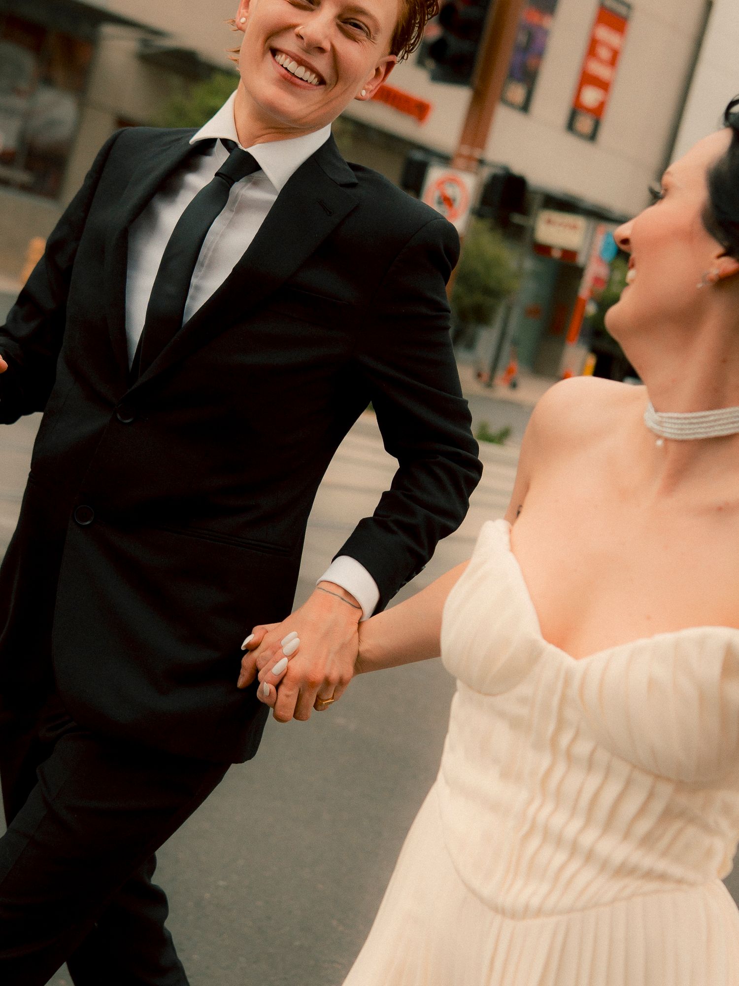 A joyful couple in wedding attire holds hands and laughs while walking together on a city street.