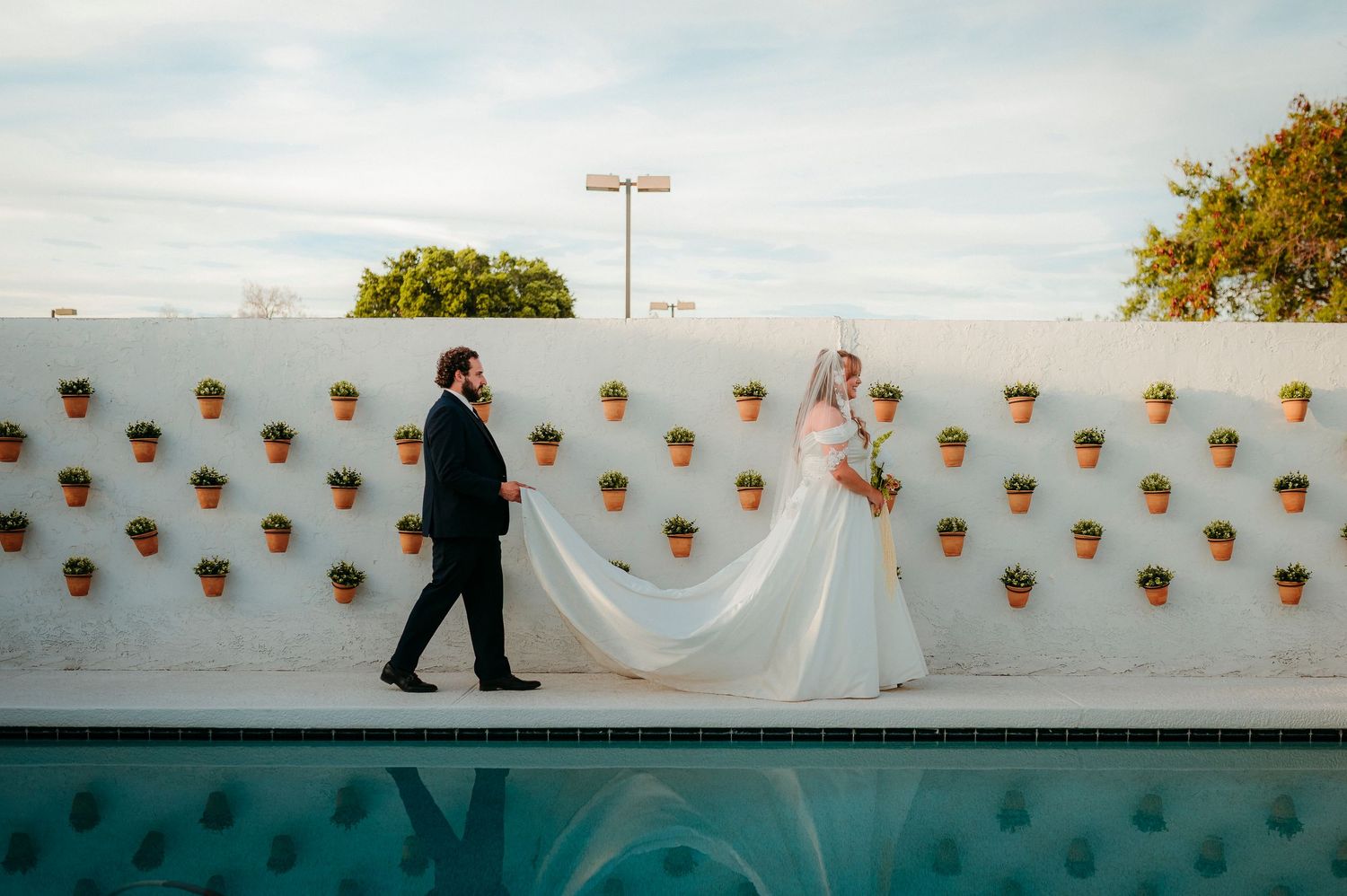 Bride and groom walk poolside past a white wall adorned with terracotta pots at an elegant outdoor wedding.