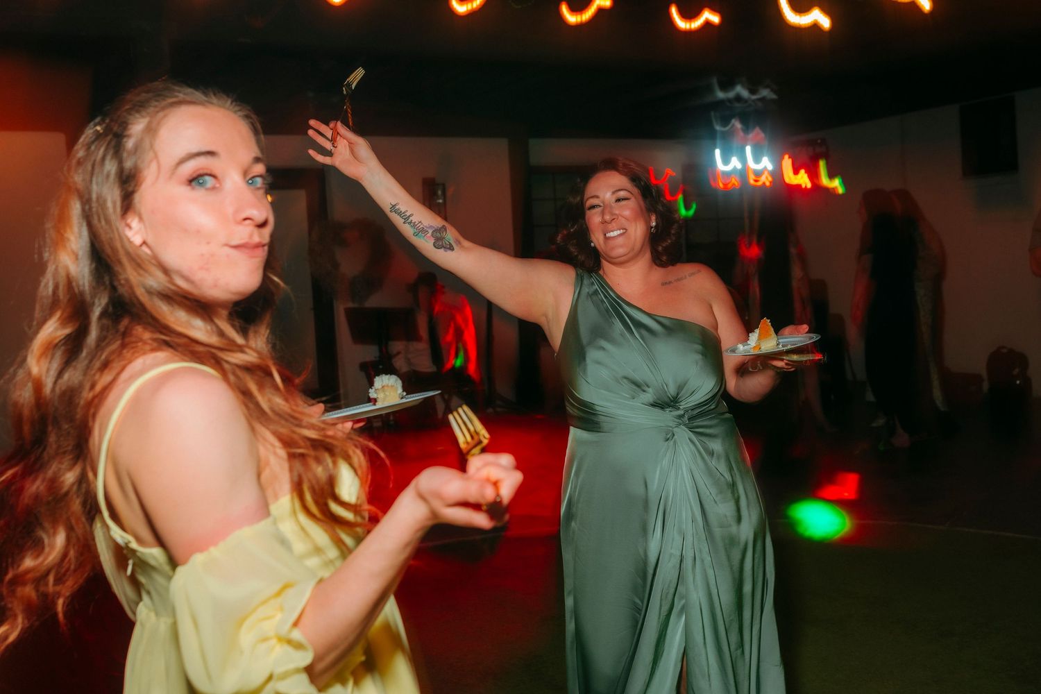 Two women in colorful bridesmaid dresses dancing and eating cake at a wedding reception with colorful lights.