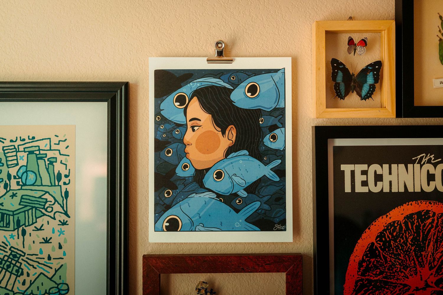 Blue illustration of woman surrounded by fish displayed in white frame on an eclectic gallery wall.