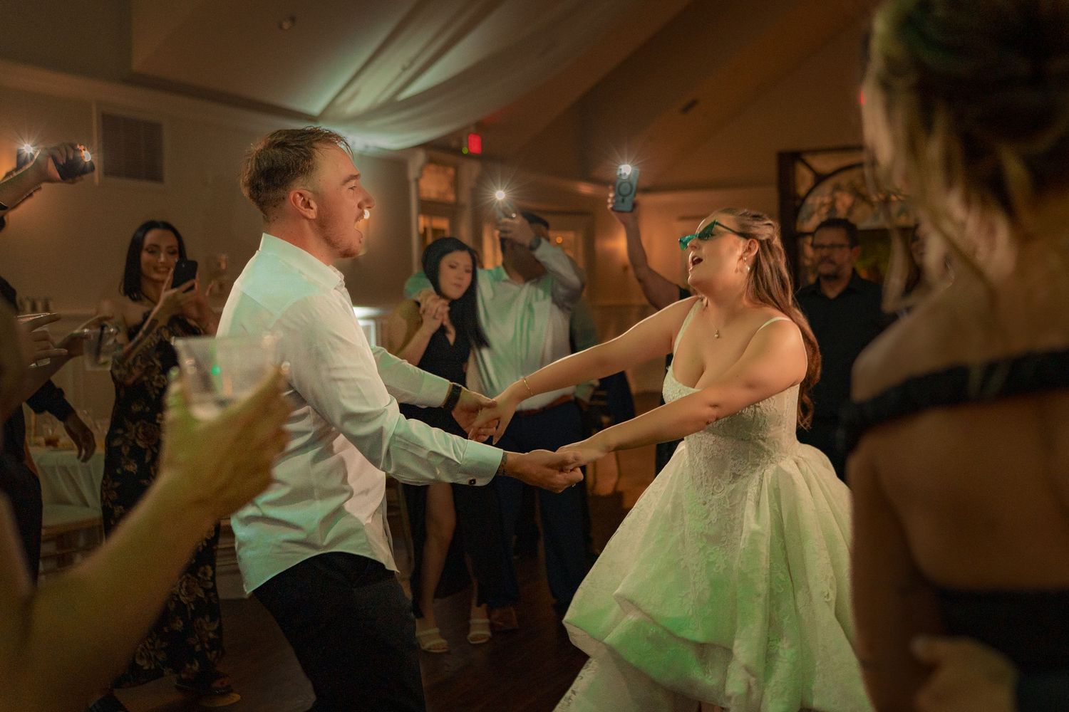 Bride and groom dance energetically together on the reception dance floor surrounded by celebrating wedding guests.