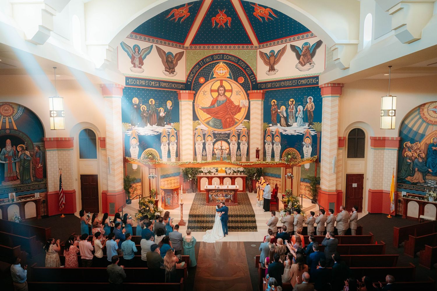 Stunning ornate church interior with colorful Byzantine murals during a wedding ceremony with guests seated in pews.