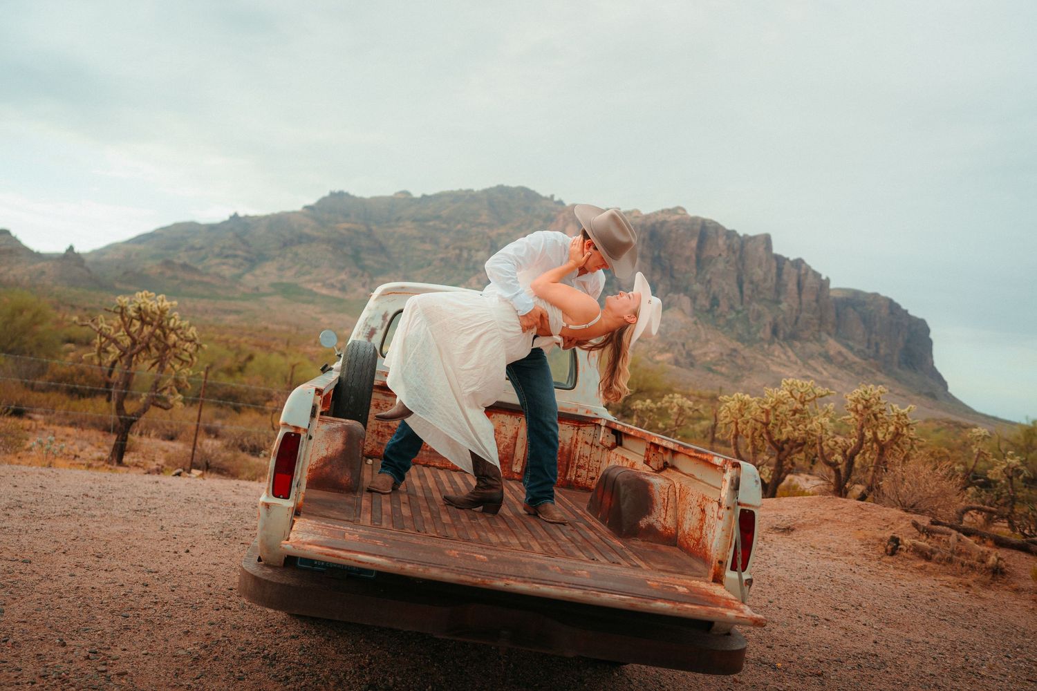 Couple sharing romantic dip kiss on rustic truck bed with Superstition Mountains backdrop in Arizona desert.