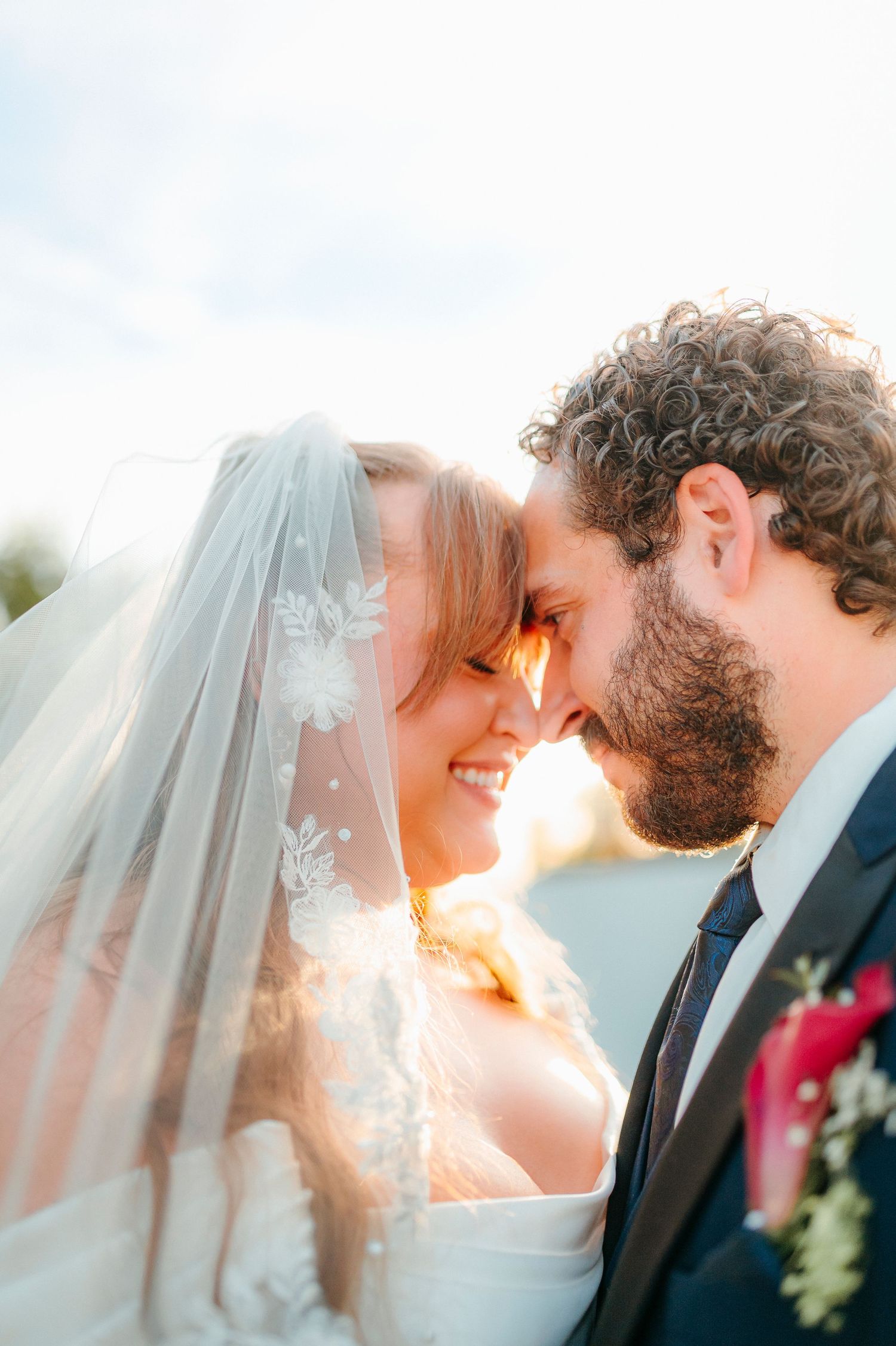 Bride and groom touching foreheads romantically, bride wearing lace veil, groom in navy suit with floral boutonniere.