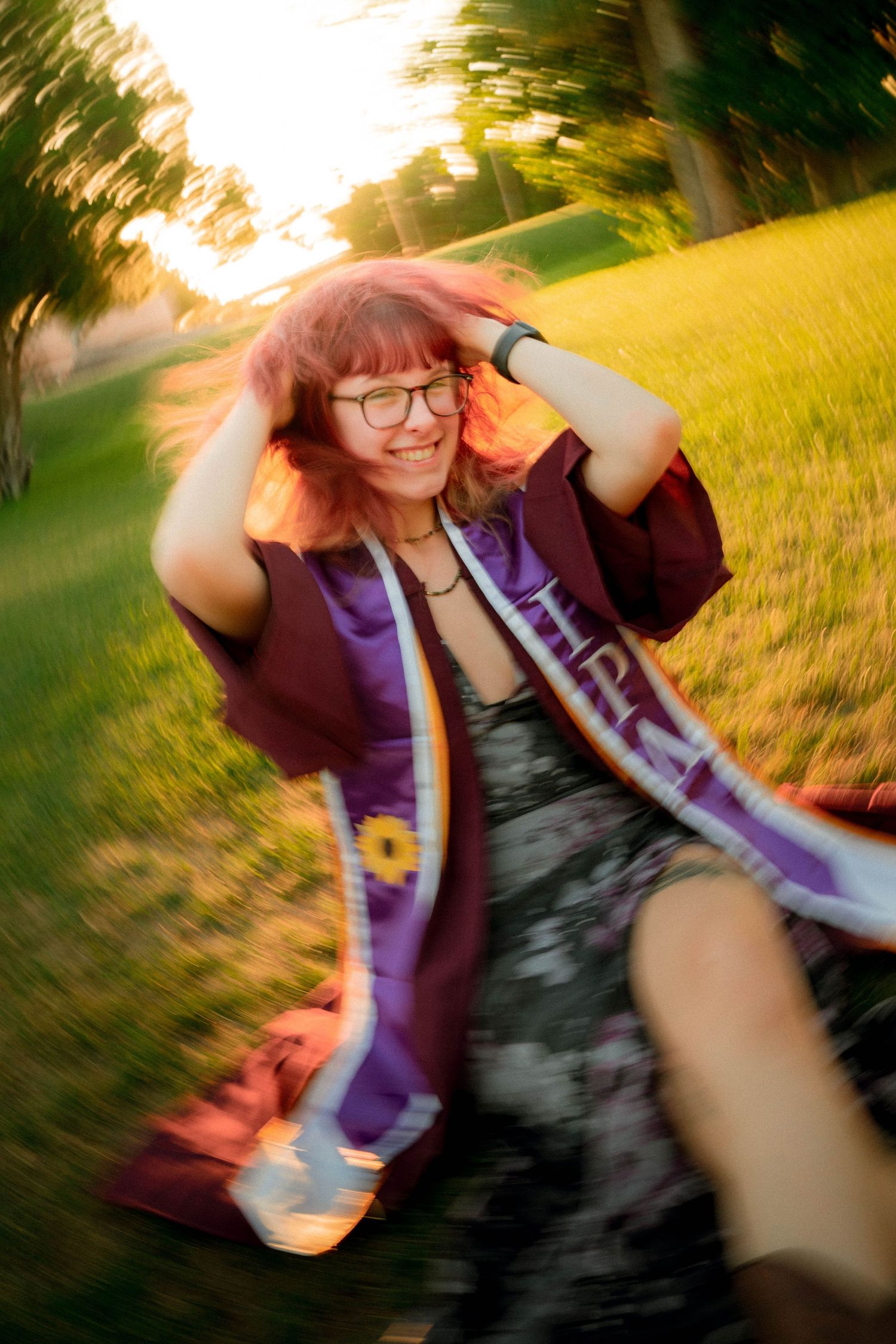Young woman in graduation regalia smiling outdoors at golden hour with motion blur effect.