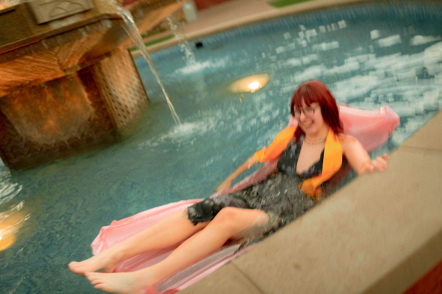 Woman with red hair relaxing on a pink float in an indoor swimming pool wearing a life vest.