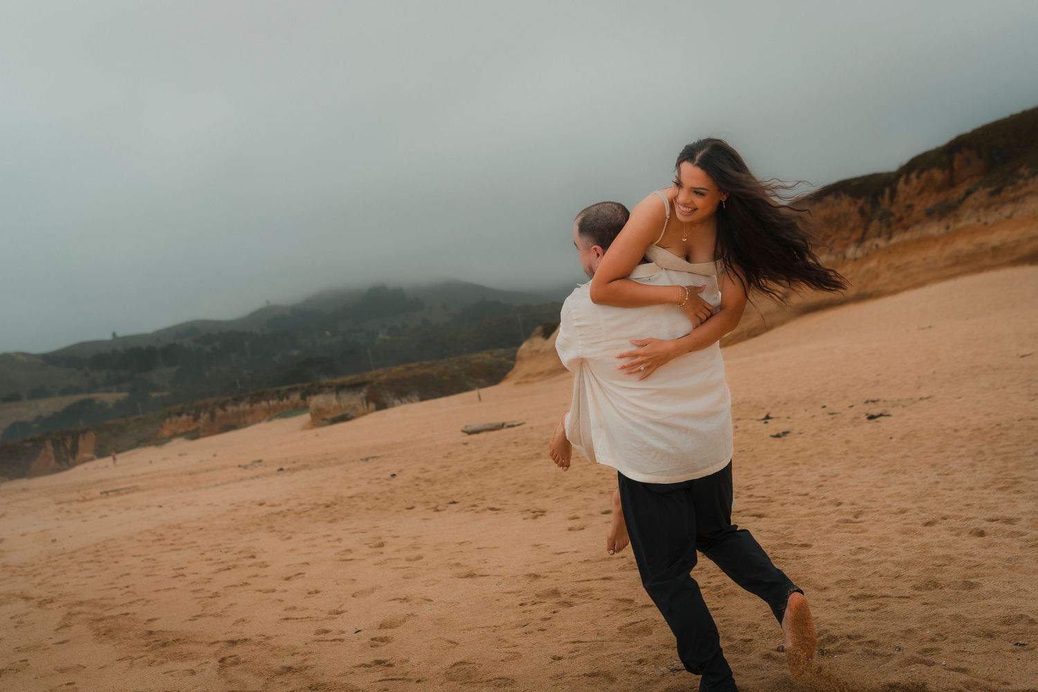 Man lifting laughing woman on a sandy beach with misty coastal cliffs in the background.